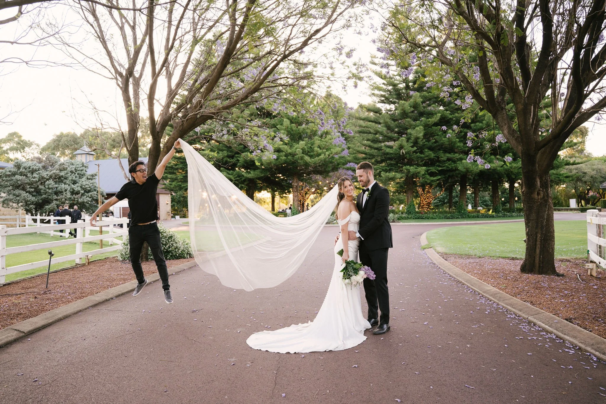 A wedding couple stands on a paved path in a park, with a woman holding up the bride's veil while smiling at the camera. The bride in a white gown holds a bouquet, and the groom in a tuxedo stands close, smiling. Trees and greenery surround them, wit