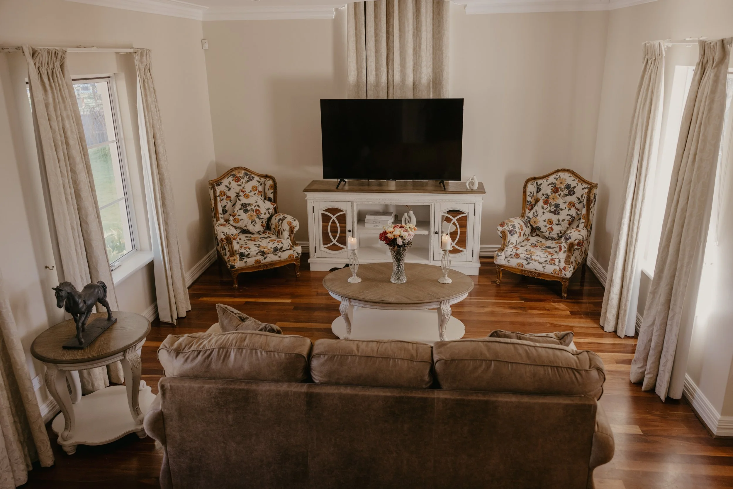 Living room with beige walls, hardwood flooring, and cream-colored curtains. There are two floral upholstered armchairs flanking a white wooden TV stand with a large flat-screen TV, decorative objects, and a flower arrangement. A beige velvet sofa is