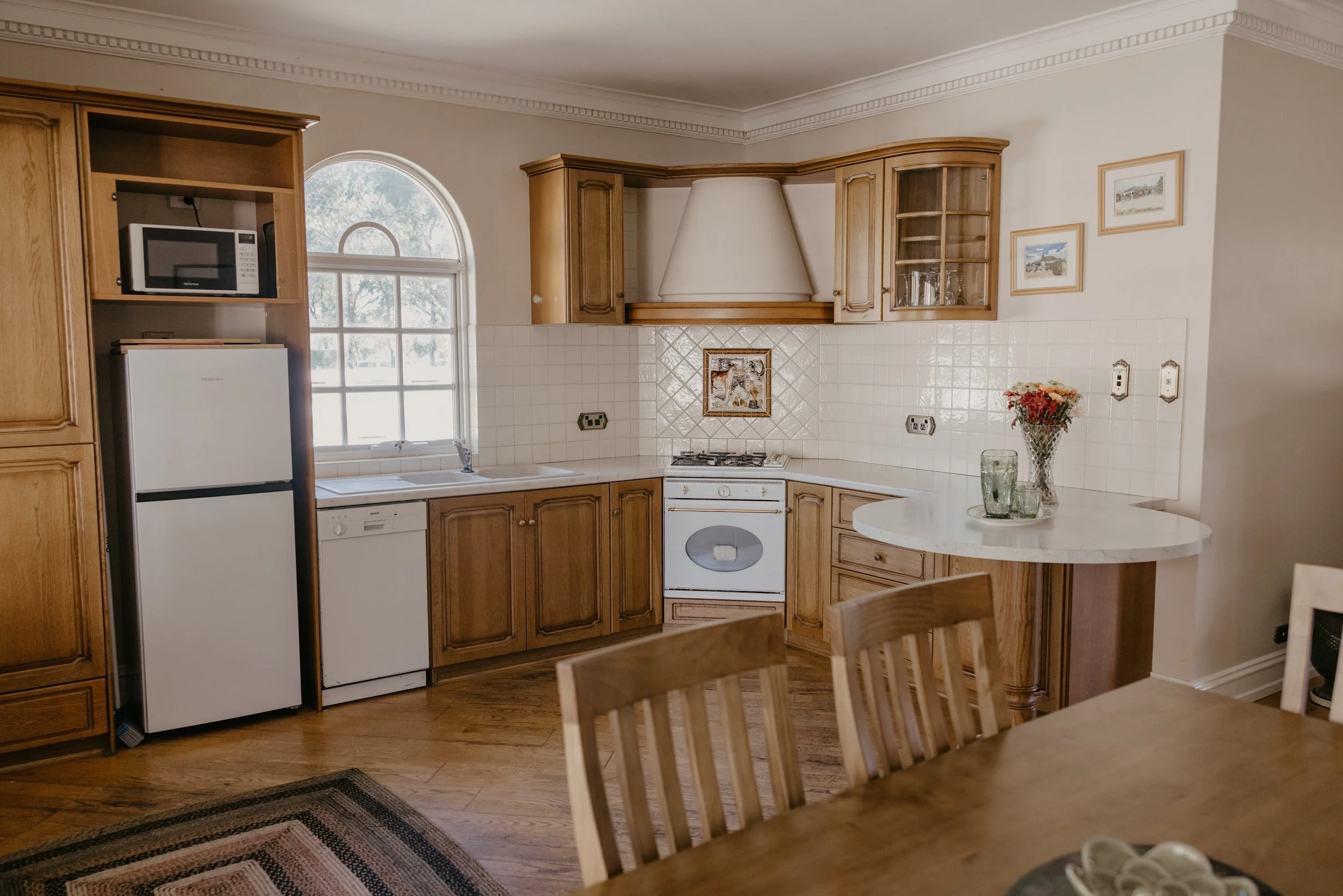 Kitchen with wooden cabinets, a white microwave on a shelf, a white refrigerator, a small oven with burners, a vase of flowers on a round counter, and framed pictures on the wall.