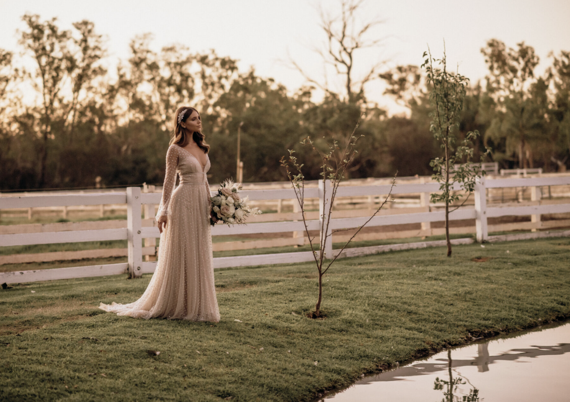 A bride in a long, white wedding gown standing outdoors near a small pond, holding a bouquet of flowers, with a white fence and trees in the background during sunset.