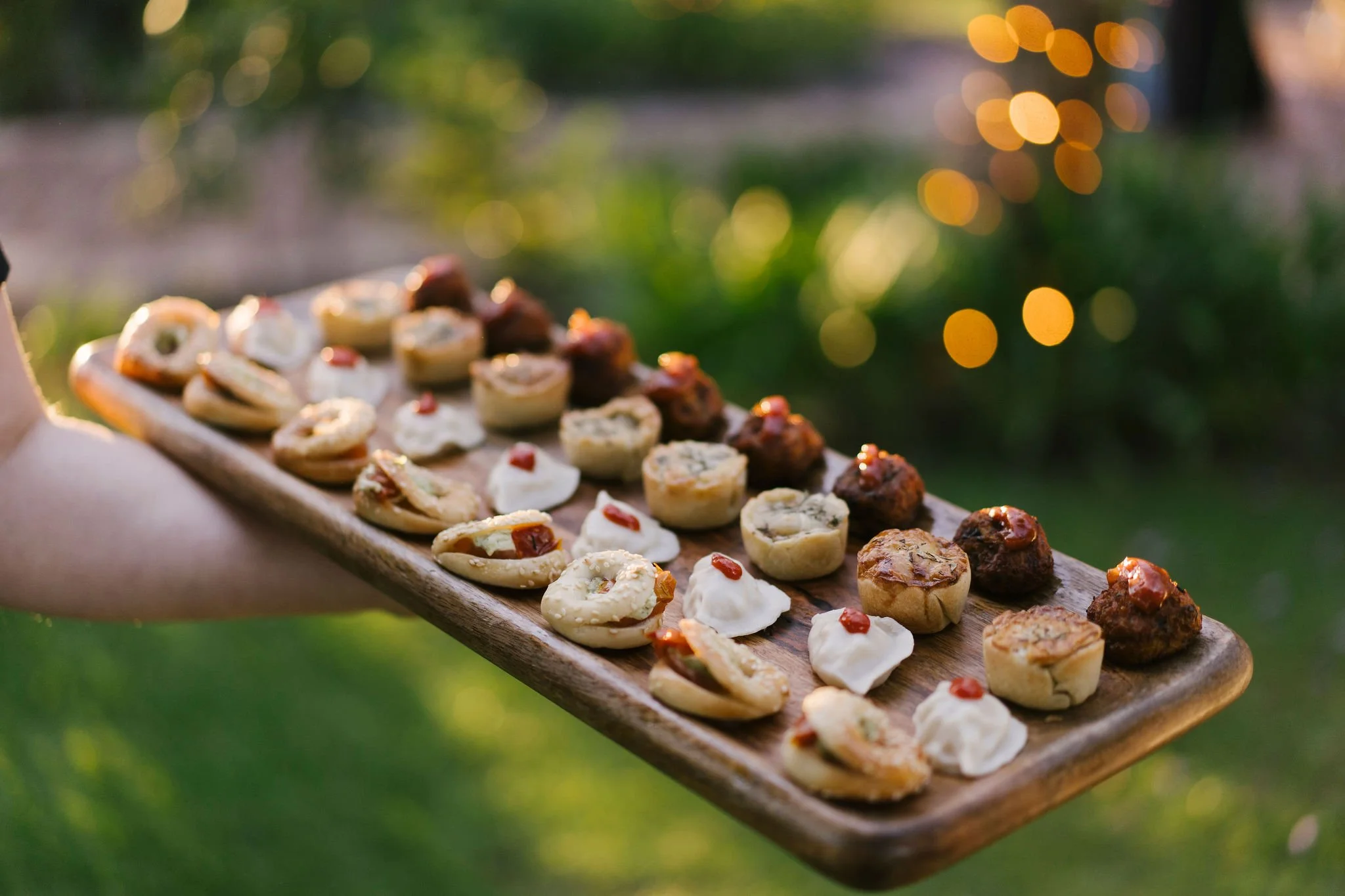 Person holding a large wooden platter of assorted small appetizers outdoors during sunset, with blurred green background and yellow bokeh lights.