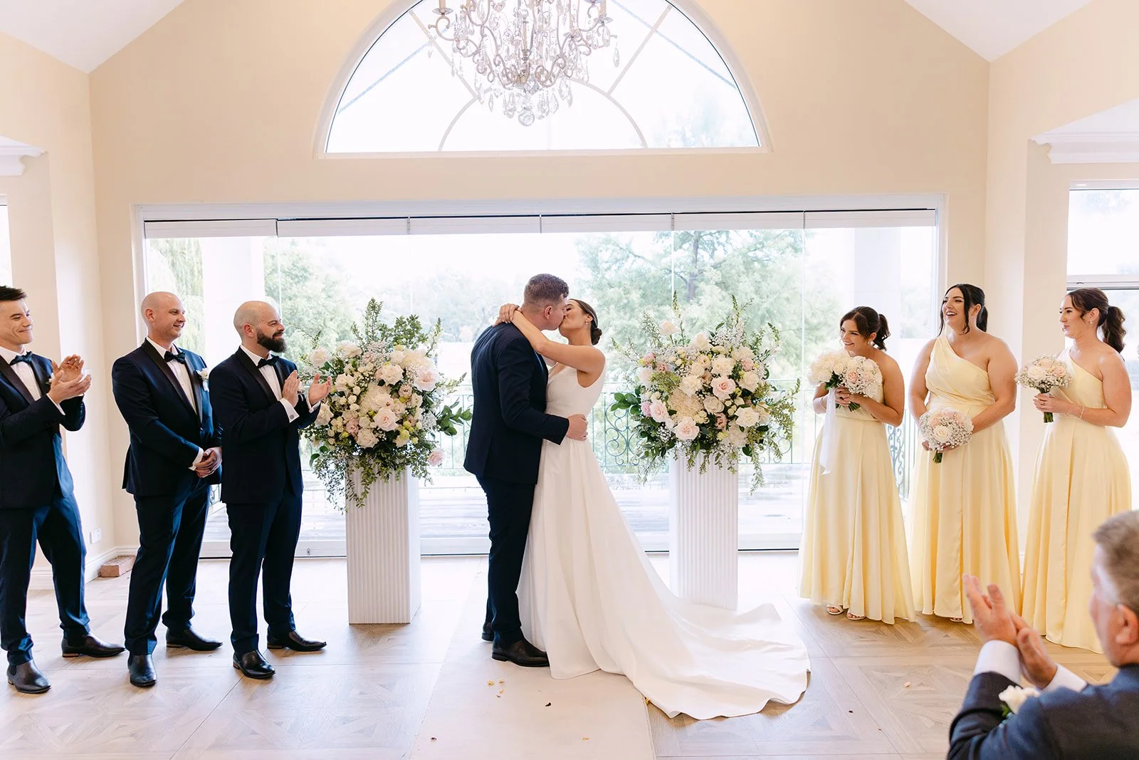 A wedding ceremony with a bride and groom kissing, surrounded by bridesmaids and groomsmen, in a bright room with large windows and floral arrangements.