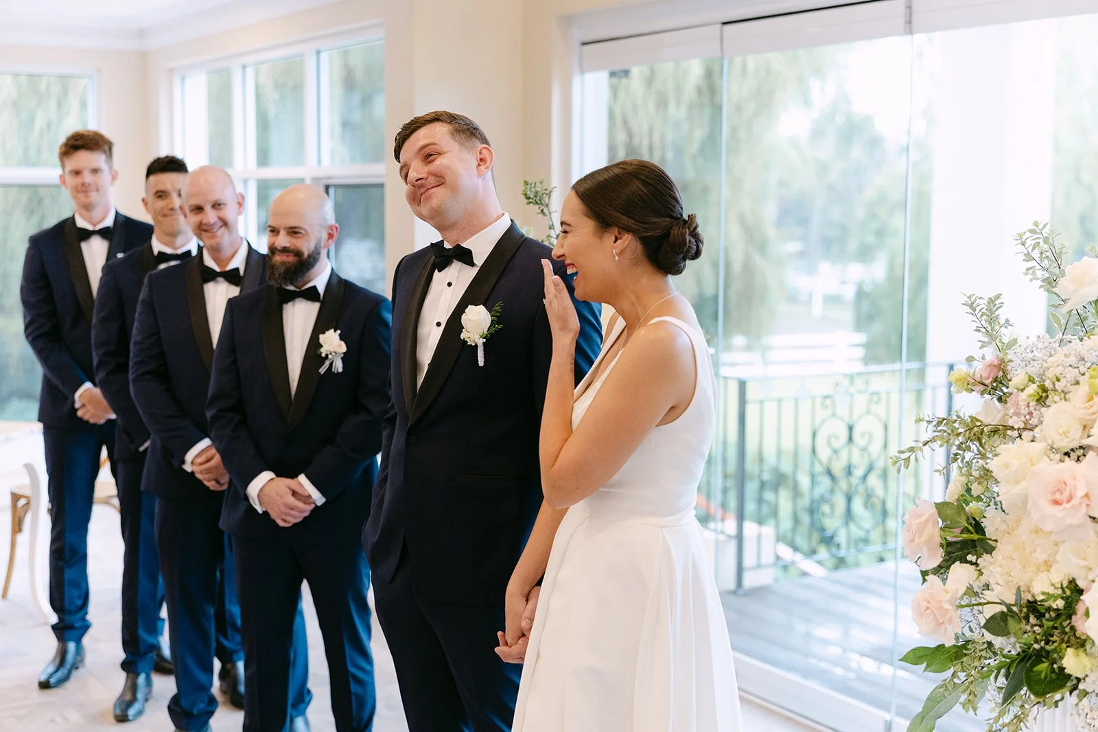 Bride and groom sharing a joyful moment during their wedding ceremony, surrounded by groomsmen and decorated with flowers in a bright room with large windows.