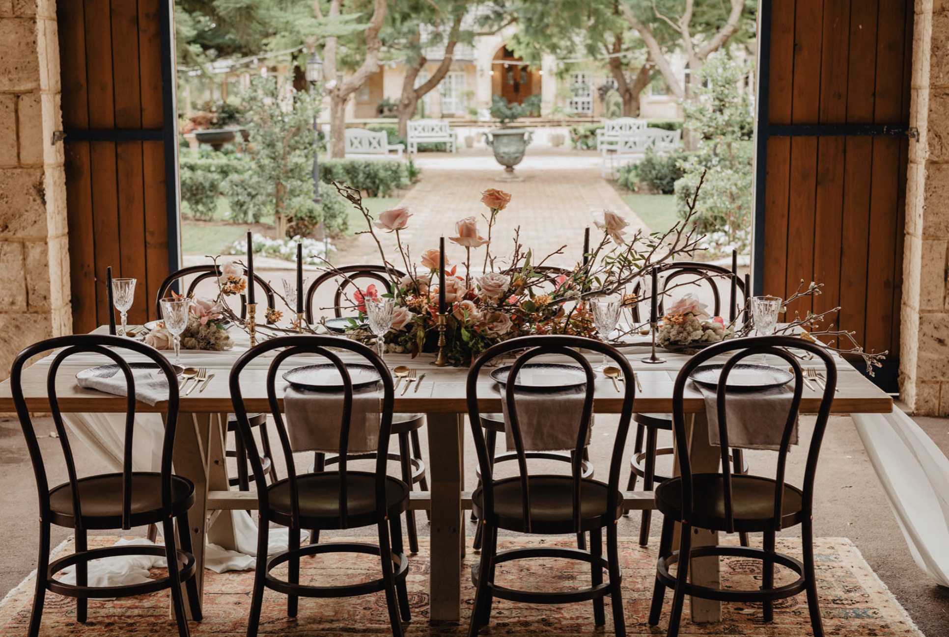 A dining table set for an event with floral centerpiece, black chairs, and elegant glassware, viewed through an open window leading to a garden with trees and benches.