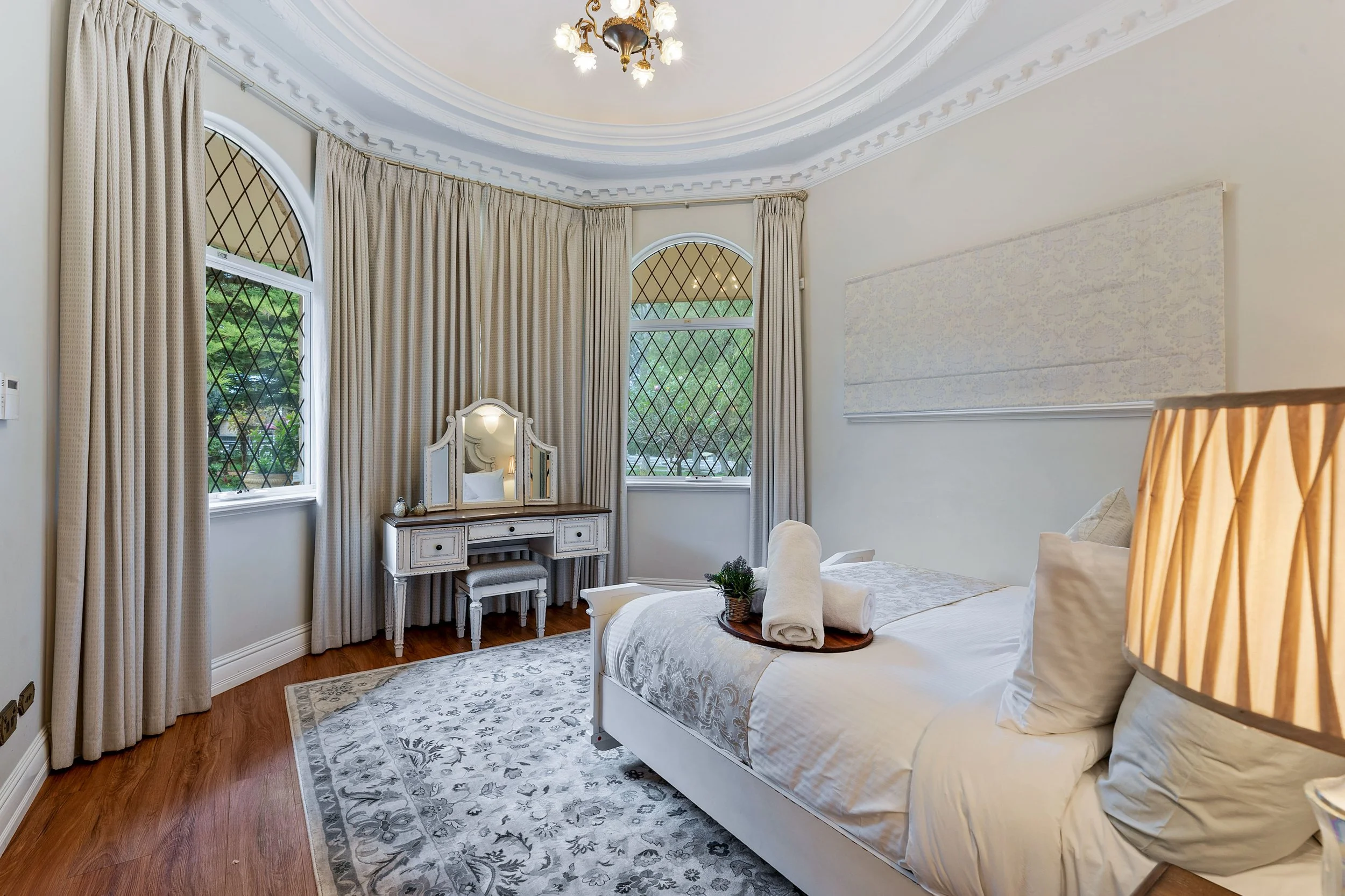 A bedroom with beige curtains covering arched windows, a white vintage vanity with mirror, a neatly made bed with rolled towels, a patterned area rug, and a chandelier hanging from a decorative ceiling.