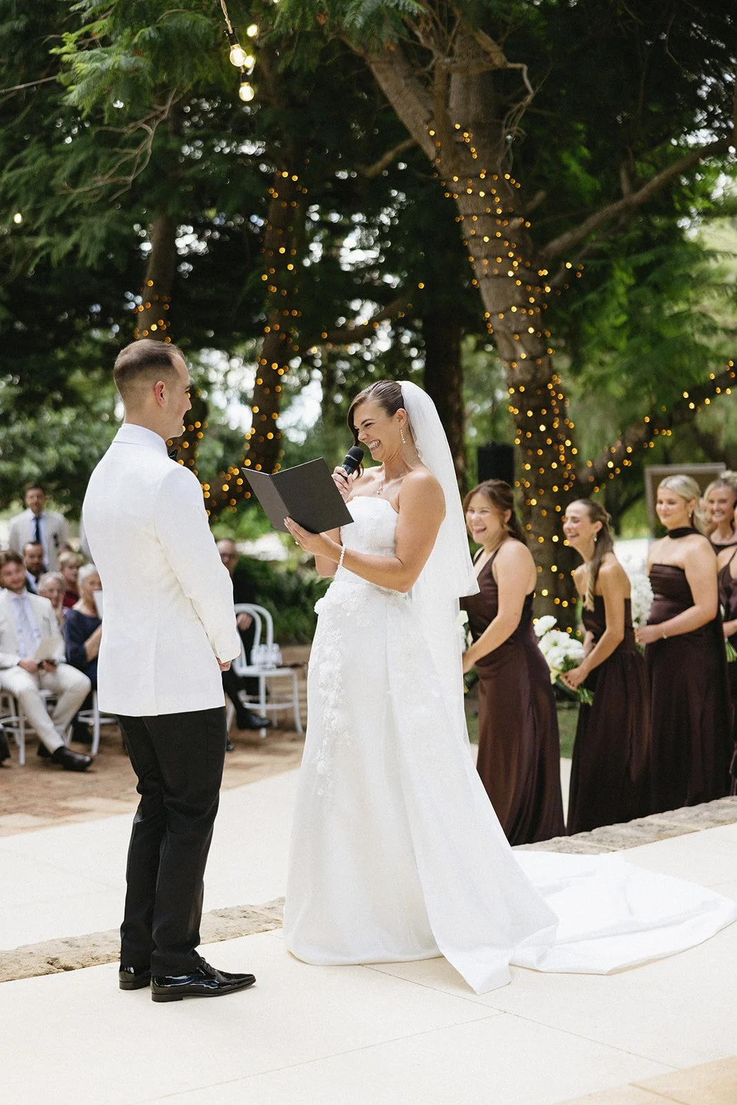 A bride and groom exchange vows during an outdoor wedding ceremony under decorated trees, with bridesmaids and guests smiling in the background.