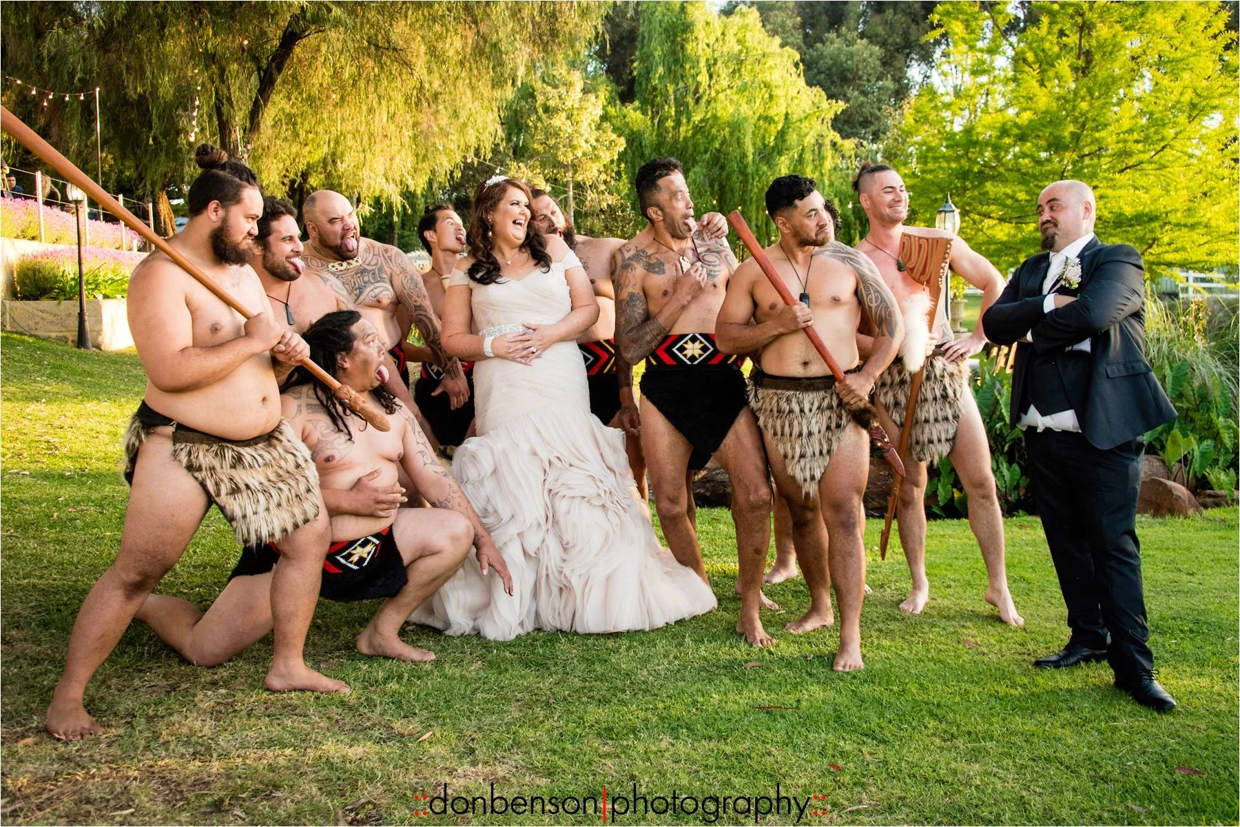 A wedding party is gathered outdoors in a lush green park with trees and landscaping, dressed in traditional Maori attire. The group, including men wearing skirts made of fur, some holding weapons, surrounds a bride in a white wedding gown laughing a