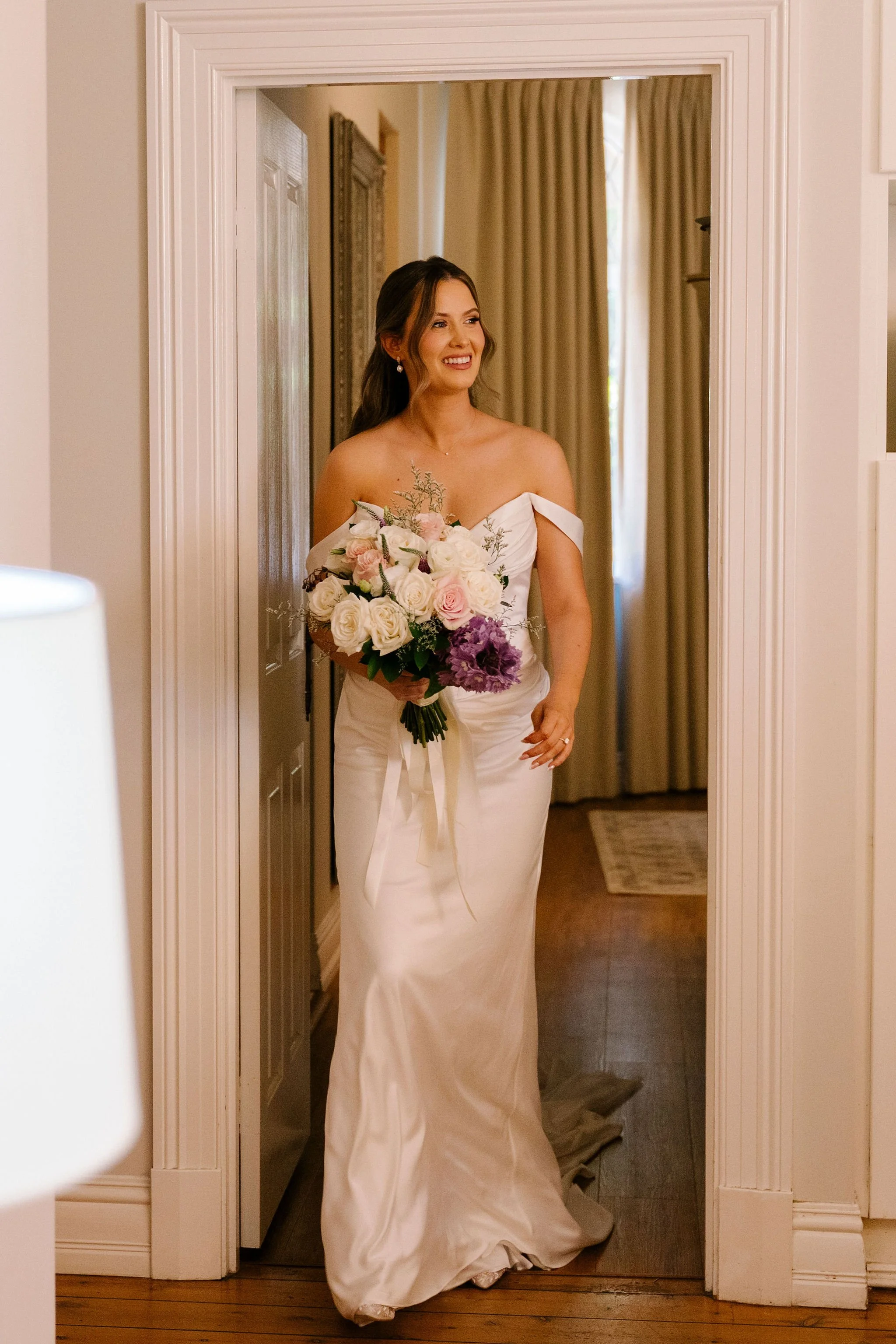 A woman in a white wedding dress holding a bouquet of white, pink, and purple flowers, smiling as she walks through a doorway in a warmly lit room.