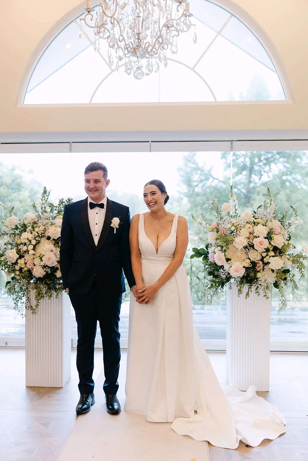 Bride and groom standing together during their wedding ceremony, smiling, with large floral arrangements on white pedestals behind them, a chandelier above, and large windows in the background.