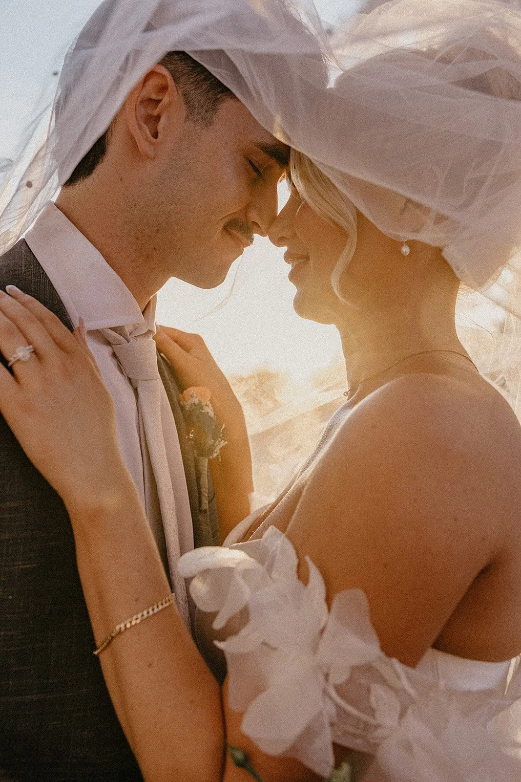 A bride and groom embrace with their foreheads touching, outdoors during sunset, both smiling gently at Brookleigh Estate Swan ValleyThe bride wears a white dress with floral details, the groom in a dark suit with a light-colored tie an boutonniere.