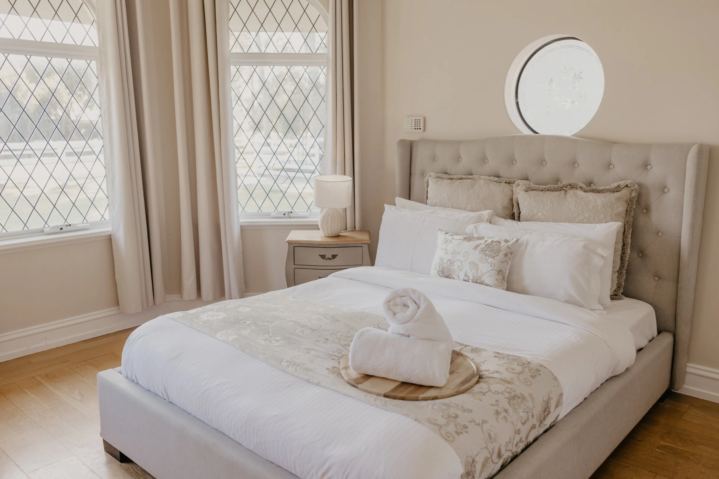 A neatly made bed with white linens and decorative pillows, a rolled towel on a wooden tray at the foot of the bed, a nightstand with a lamp, and large windows with diamond-patterned panes and cream curtains in a bright bedroom.