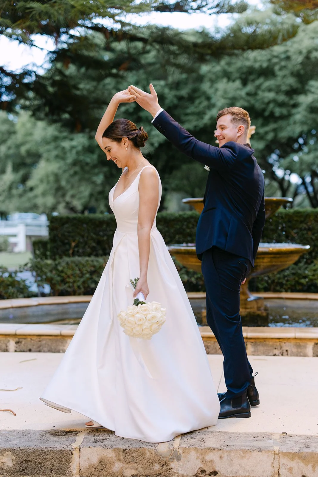 A newlywed couple dancing outdoors by a fountain. The bride is holding a bouquet of white roses, and the groom is twirling her with a smile.