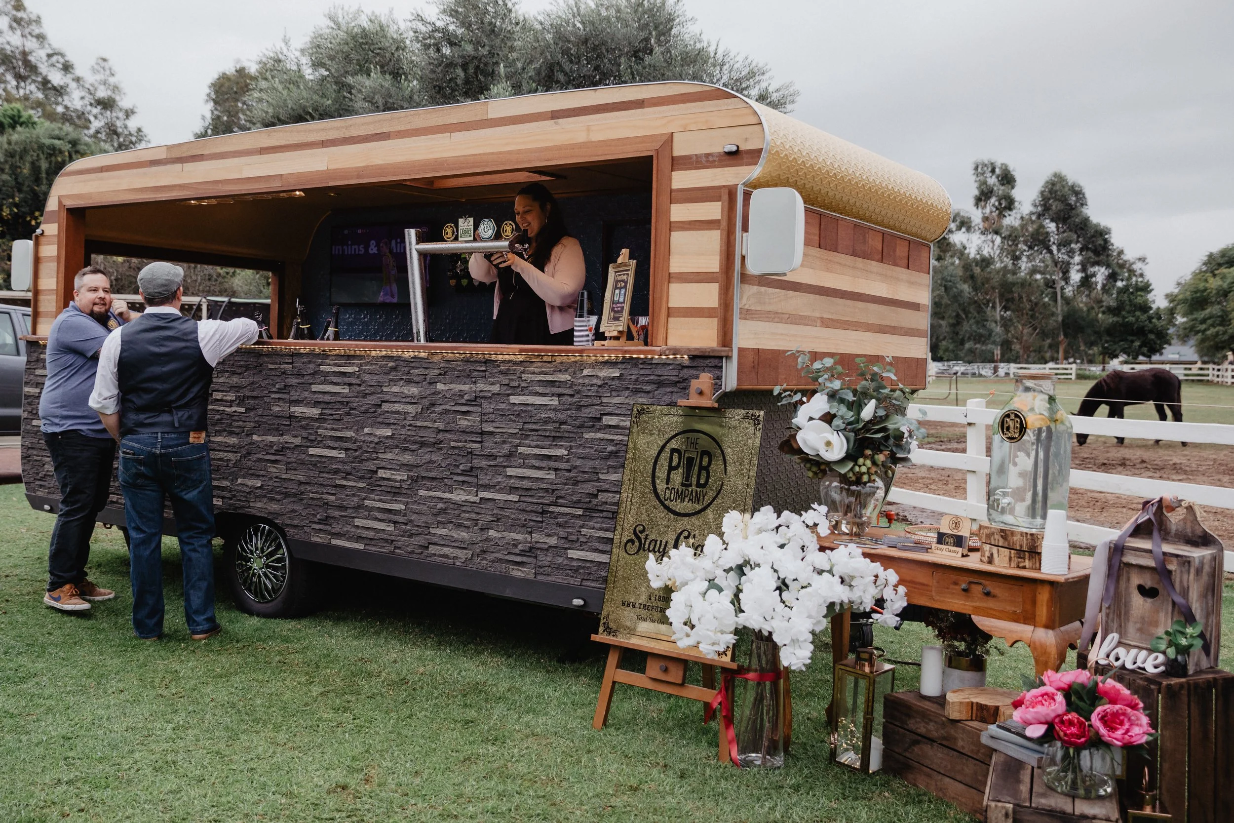 A mobile bar with a wood exterior serving drinks at an outdoor event, with two men ordering, and a woman bartender behind the counter. Decor includes flowers and a sign reading "Stay Classy."