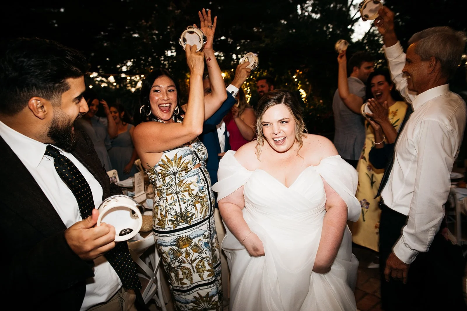 Group of people celebrating at a wedding reception, with the bride in a white dress in the center, smiling and dancing. Others around her are cheering and holding tambourines, enjoying the festive moment.