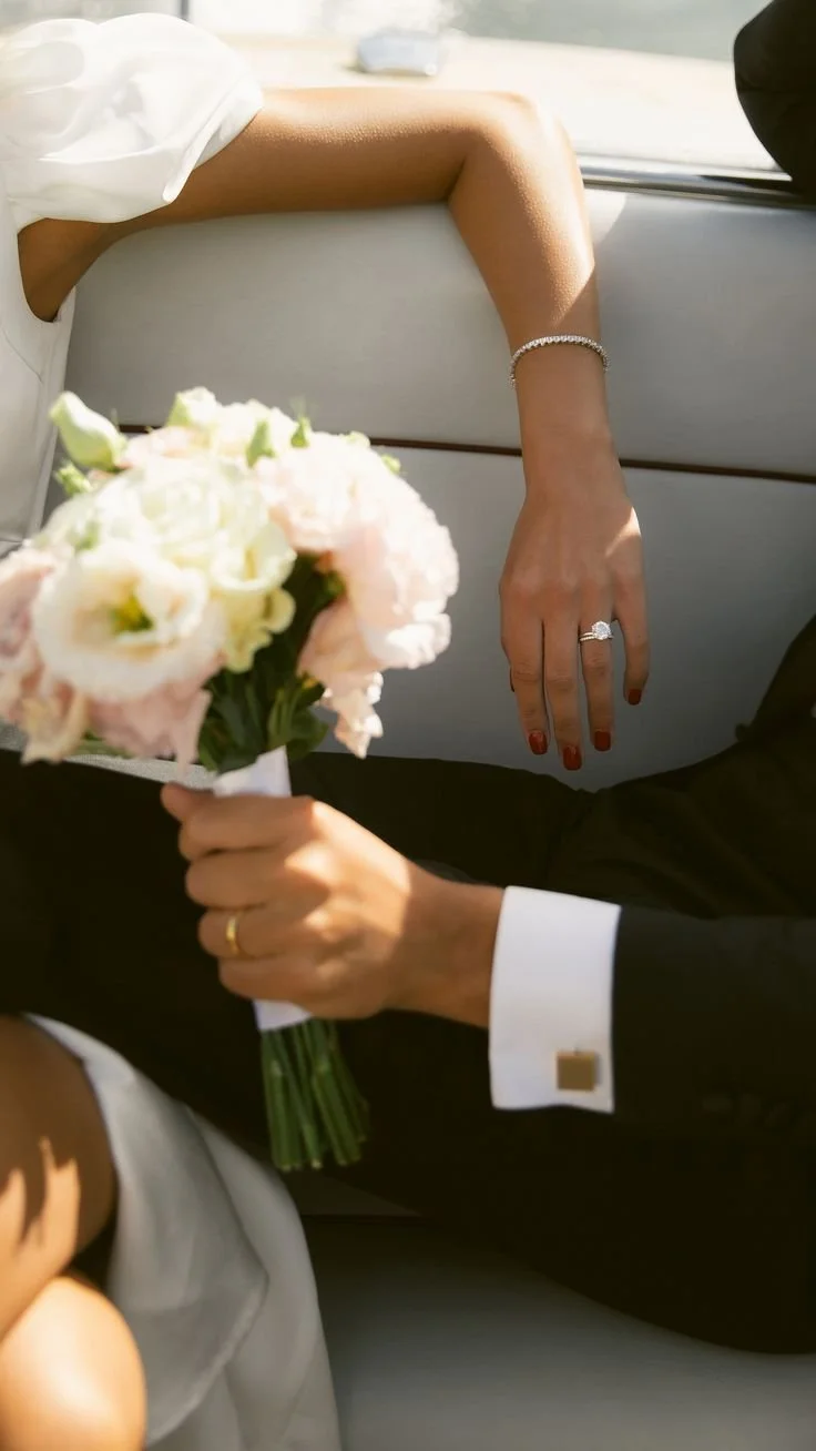 A bride and groom sitting in a car, with the bride holding a bouquet of flowers. The bride's hand, with an engagement ring, rests on the car seat, and she is wearing a silver bracelet. The groom is in a black suit with white shirt cuffs.