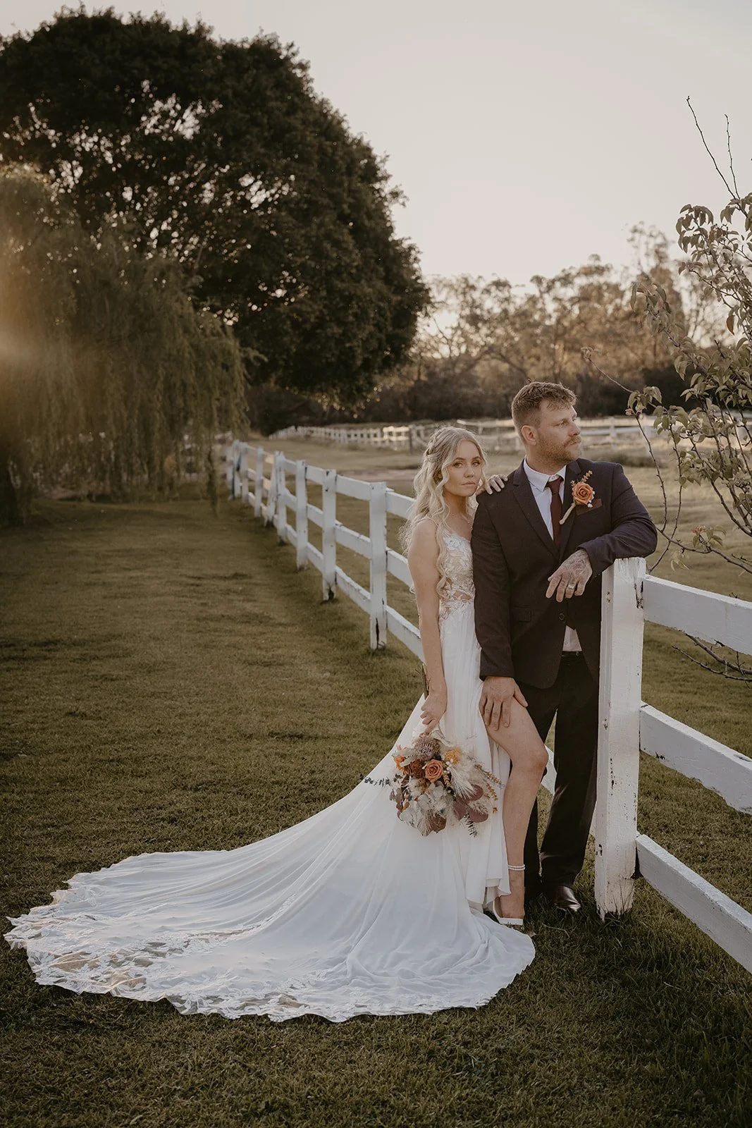 A bride and groom standing outdoors near a white wooden fence during sunset, with trees in the background. The bride is wearing a white wedding dress with a long train, holding a bouquet, and the groom is wearing a dark suit with a boutonniere.