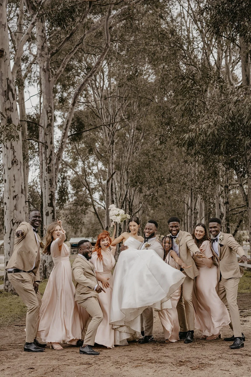 Smiling wedding party in beige suits and blush bridesmaids' dresses, with the bride and groom in the center, outdoors among tall trees.