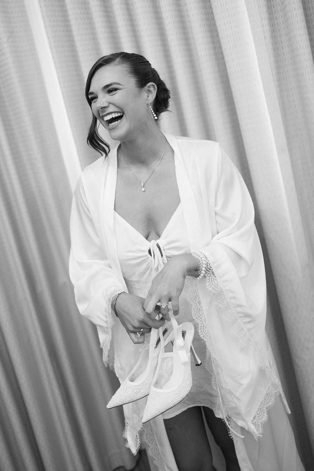 A woman with short dark hair, dressed in white, is smiling and holding a pair of white high-heeled shoes. She is standing in front of a curtain, wearing jewelry and a loose top with lace details.