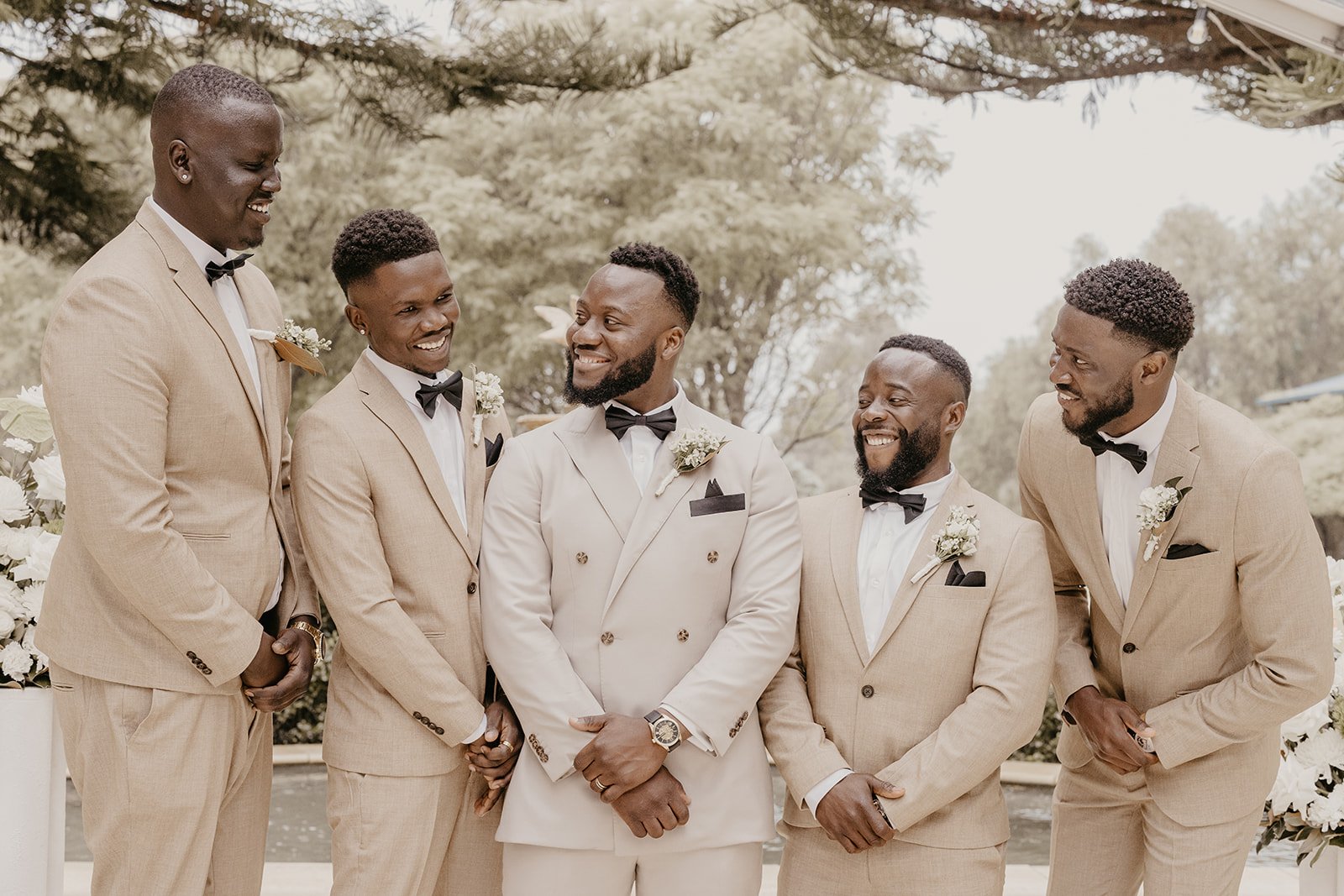 A group of six men in beige suits and black bow ties, standing outdoors, smiling and looking at each other, during a wedding celebration.