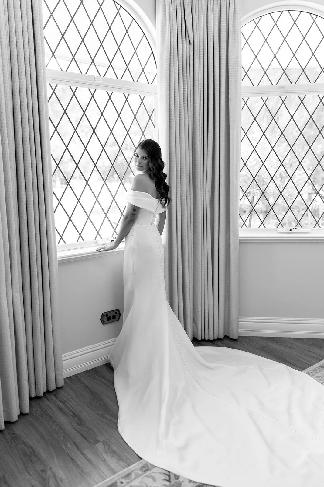 A woman in an off-the-shoulder white wedding gown standing by a window with diamond-patterned leaded glass, curtains drawn to the side, in a bright room with wooden flooring.