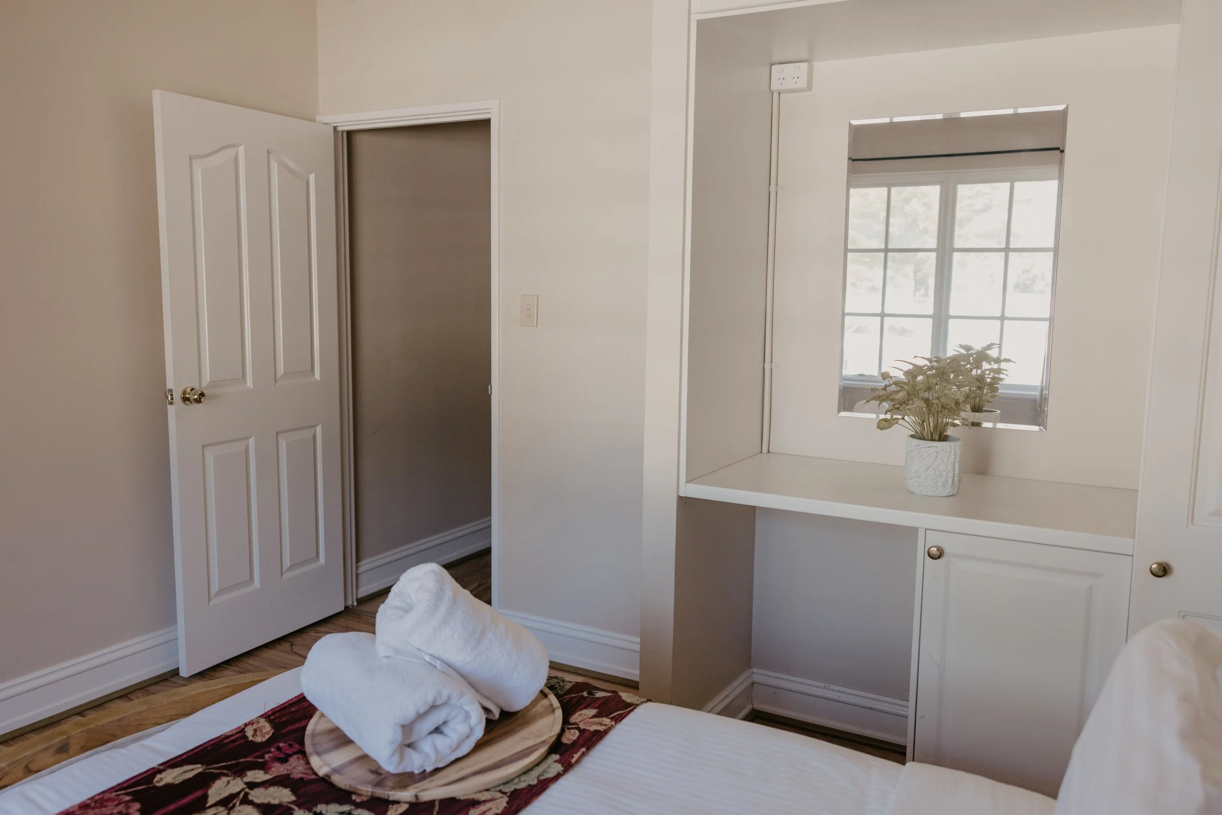 A cozy bedroom with a white wall, a mirror, a window with sunlight, and a potted plant. Towels are folded on a tray at the foot of the bed.