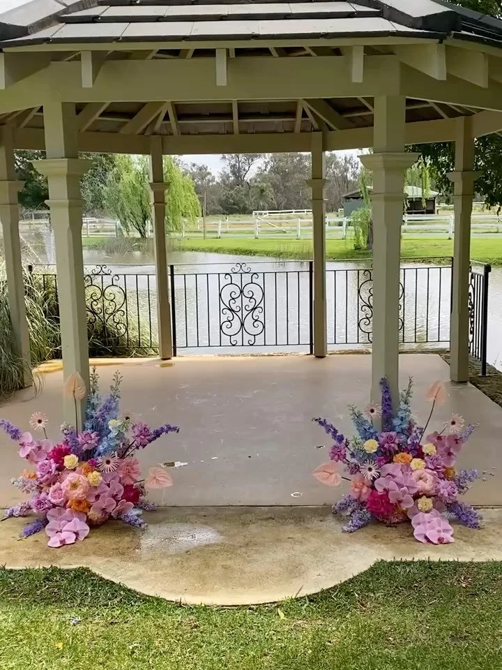 Pink and purple floral arrangements at the front of a gazebo overlooking a pond with a fence, trees, and a building in the background.