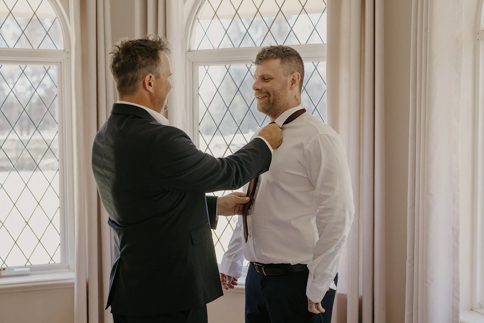 Two men in formal attire are standing by a large window with diamond-shaped panes. One man, in a dark suit, is helping the other man, in a white dress shirt and dark pants, adjust his black tie. The scene suggests a wedding or special occasion prepar