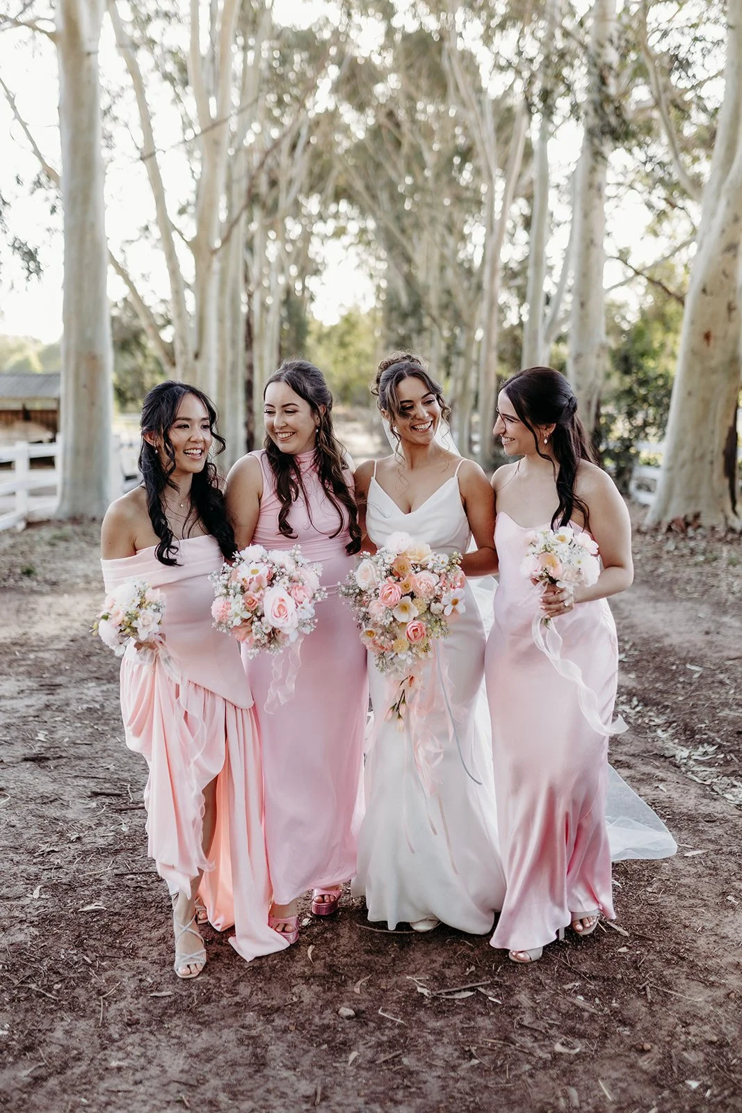 Four women in bridesmaid dresses standing outdoors amongst trees, holding bouquets, smiling and looking at each other.