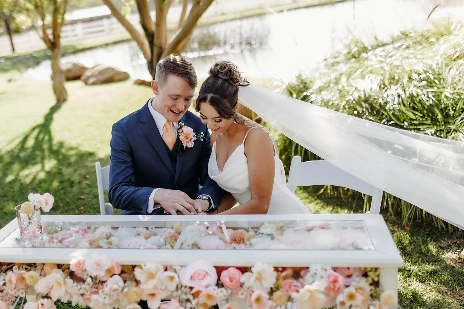A bride and groom sit together outdoors during their wedding, looking at their hands, surrounded by flowers and greenery.