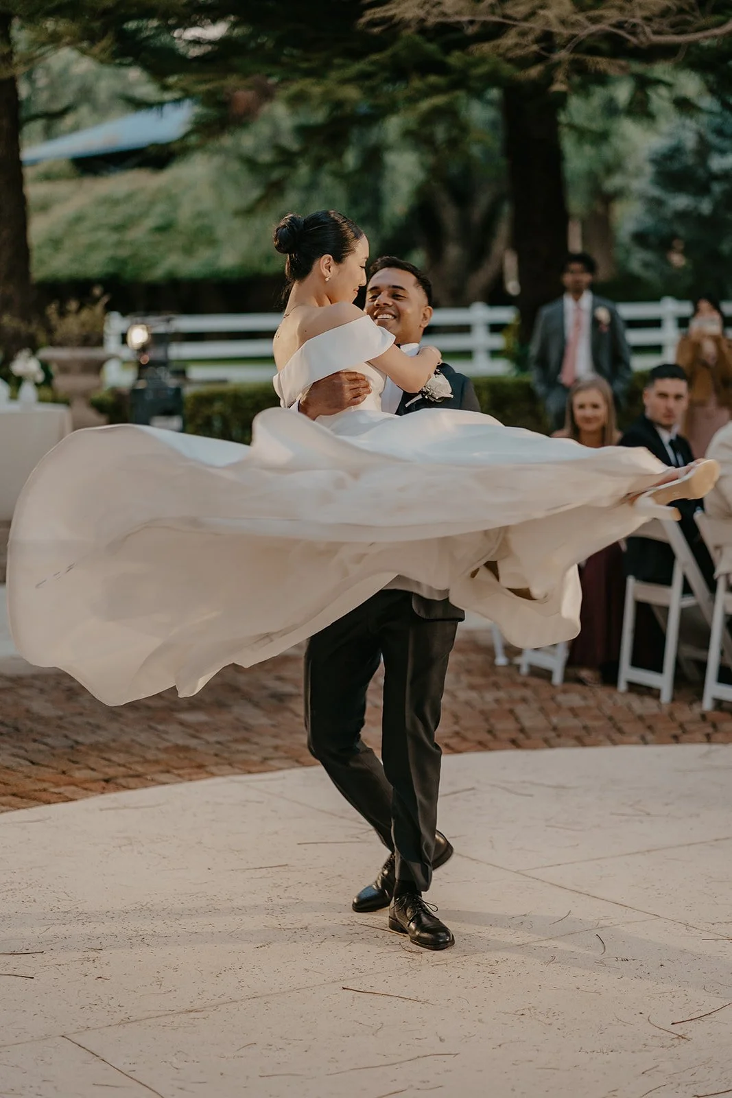 Groom lifting bride while dancing at an outdoor wedding reception surrounded by guests and greenery.