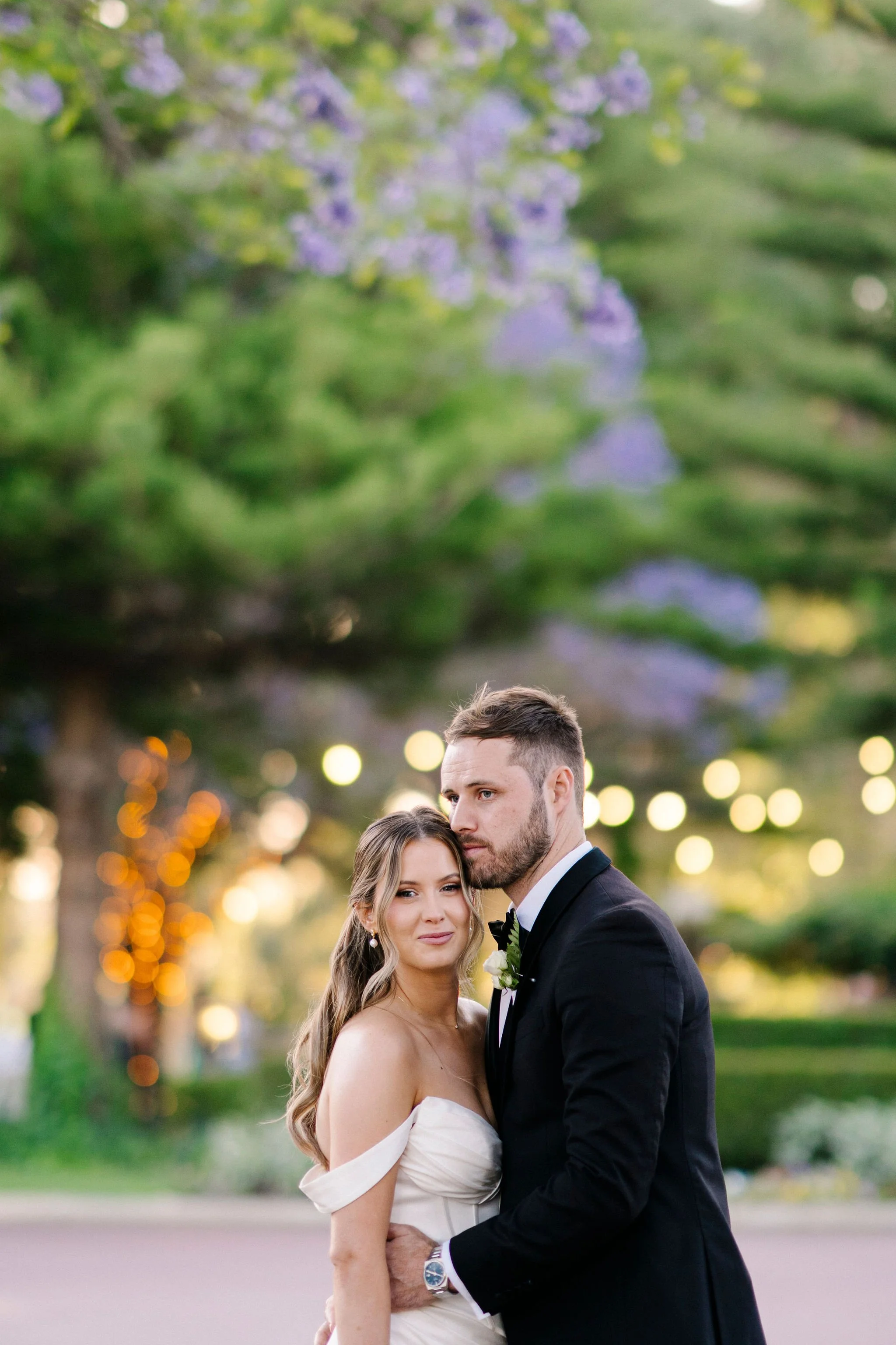 A bride and groom pose outdoors on their wedding day, with blurred trees and string lights in the background.