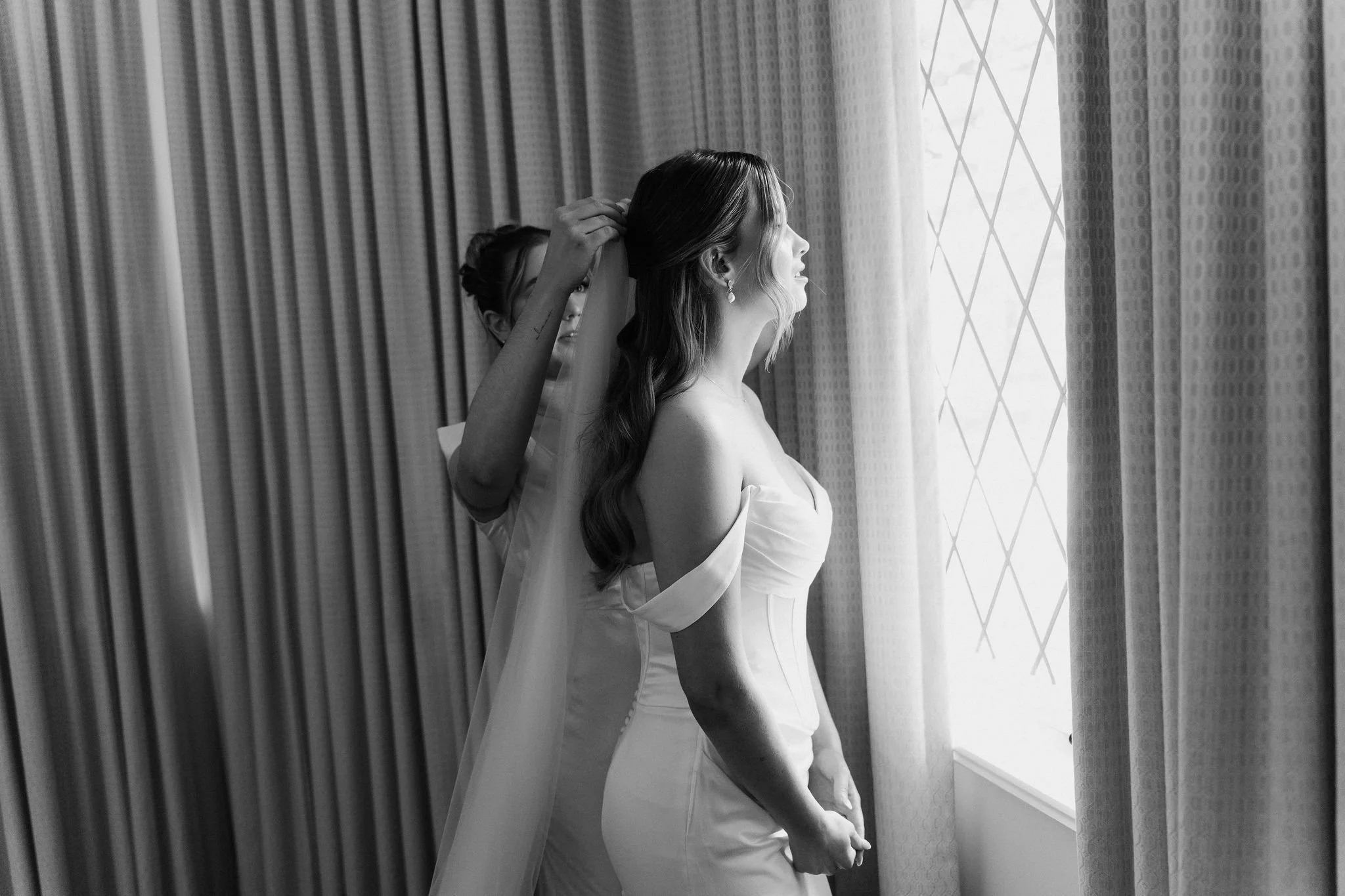 A bride with wavy hair looking out a window as someone helps her with her veil.