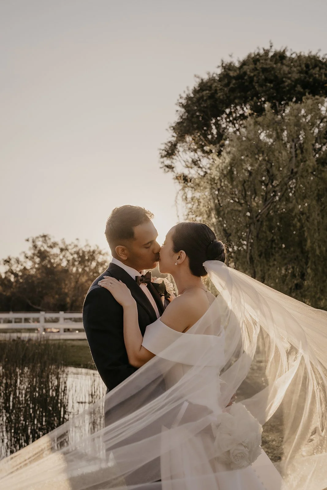 A newlywed couple sharing a kiss outdoors at sunset, with the bride in a wedding dress and veil, and the groom in a tuxedo, near a pond and trees.