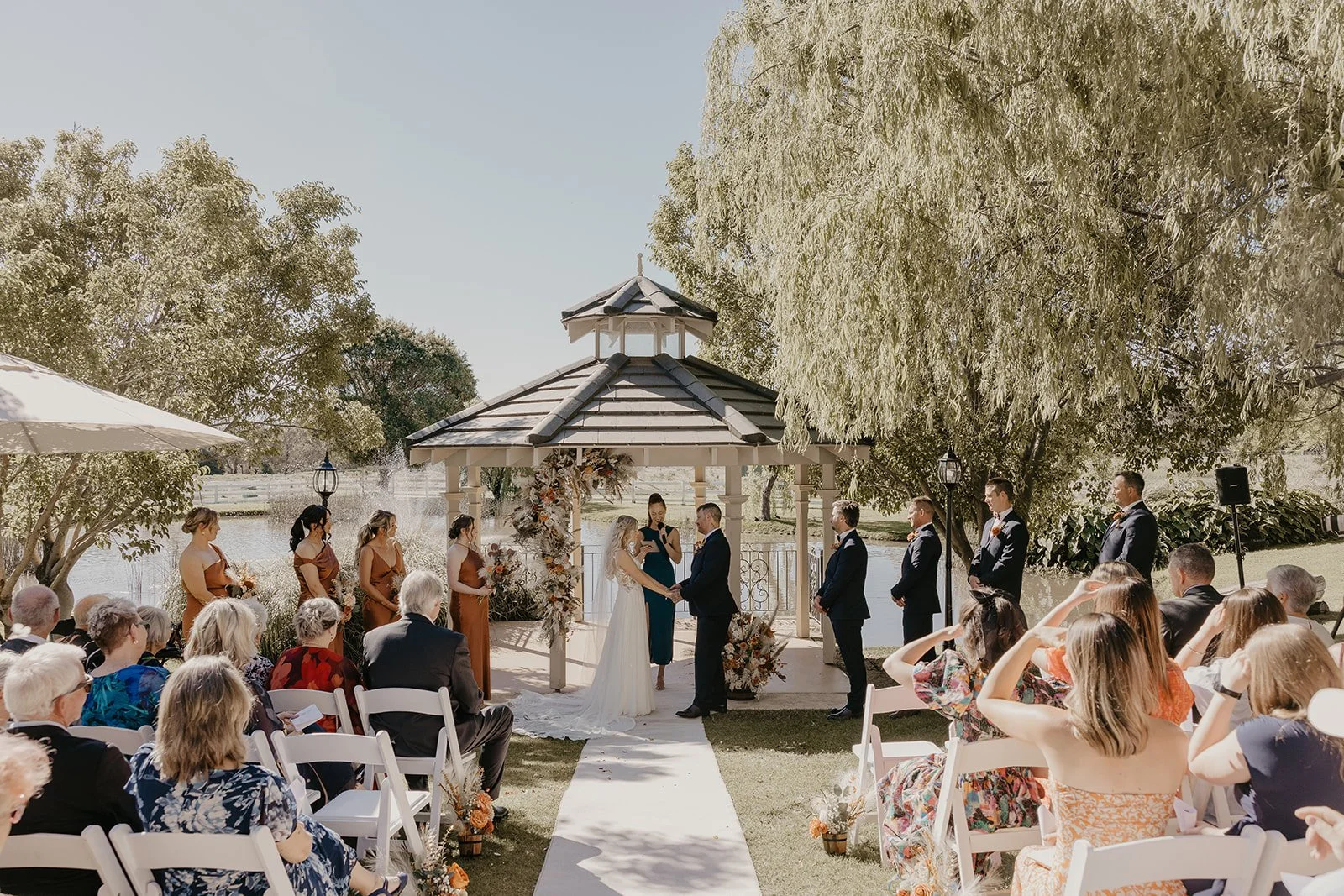 Outdoor wedding ceremony taking place under a gazebo by a lake with guests seated on white chairs and a large tree to the right.
