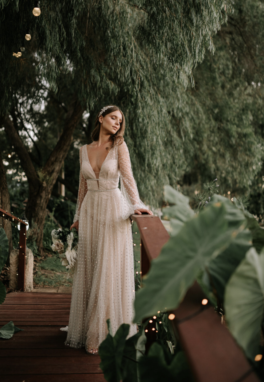 A bride in a white wedding dress with long sheer sleeves and a deep V neckline, holding a bouquet, standing on a wooden bridge decorated with string lights, surrounded by lush tall green trees and plants.