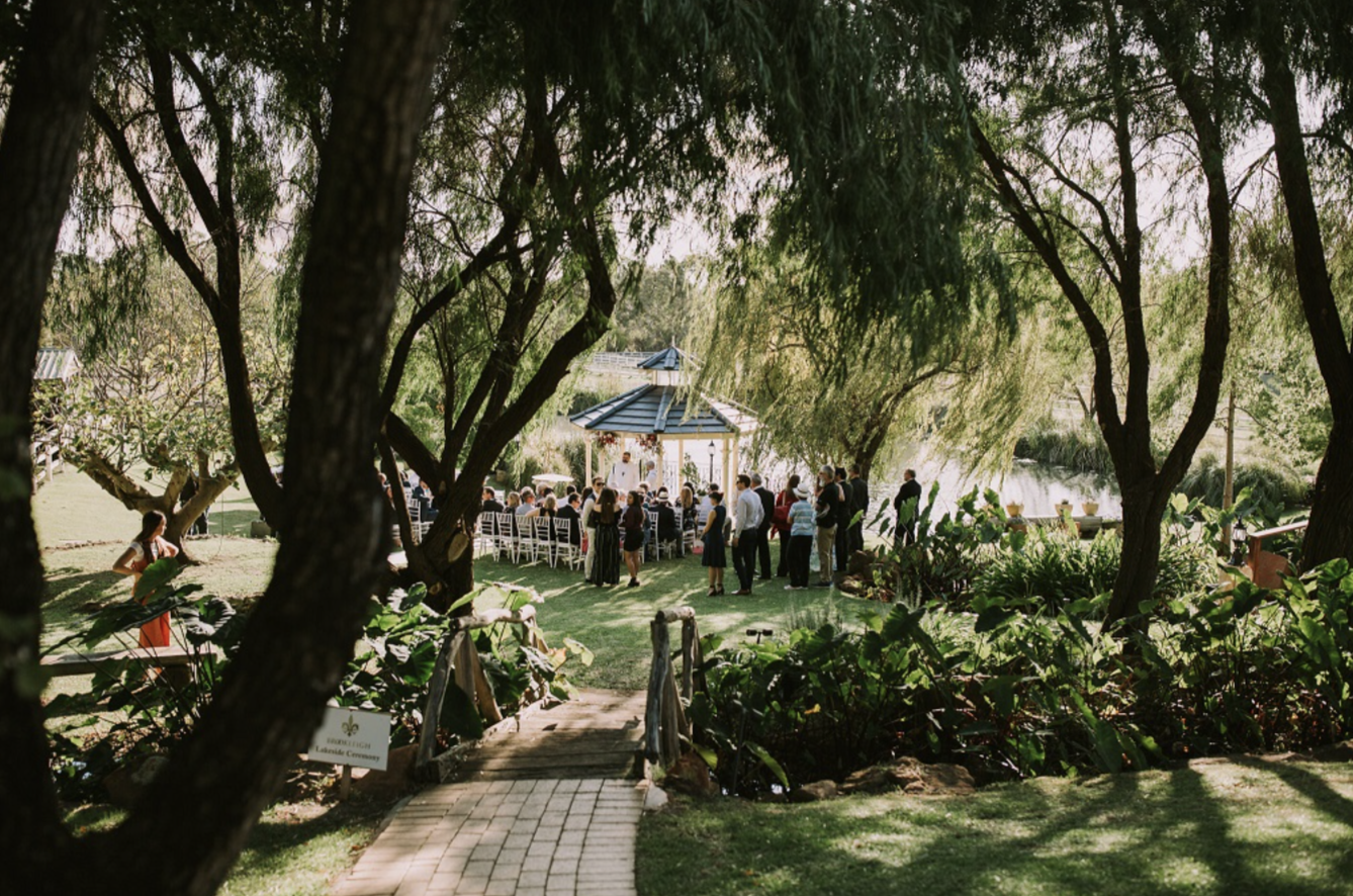 Outdoor wedding ceremony under trees with guests seated at tables and standing, near a gazebo by a lake.