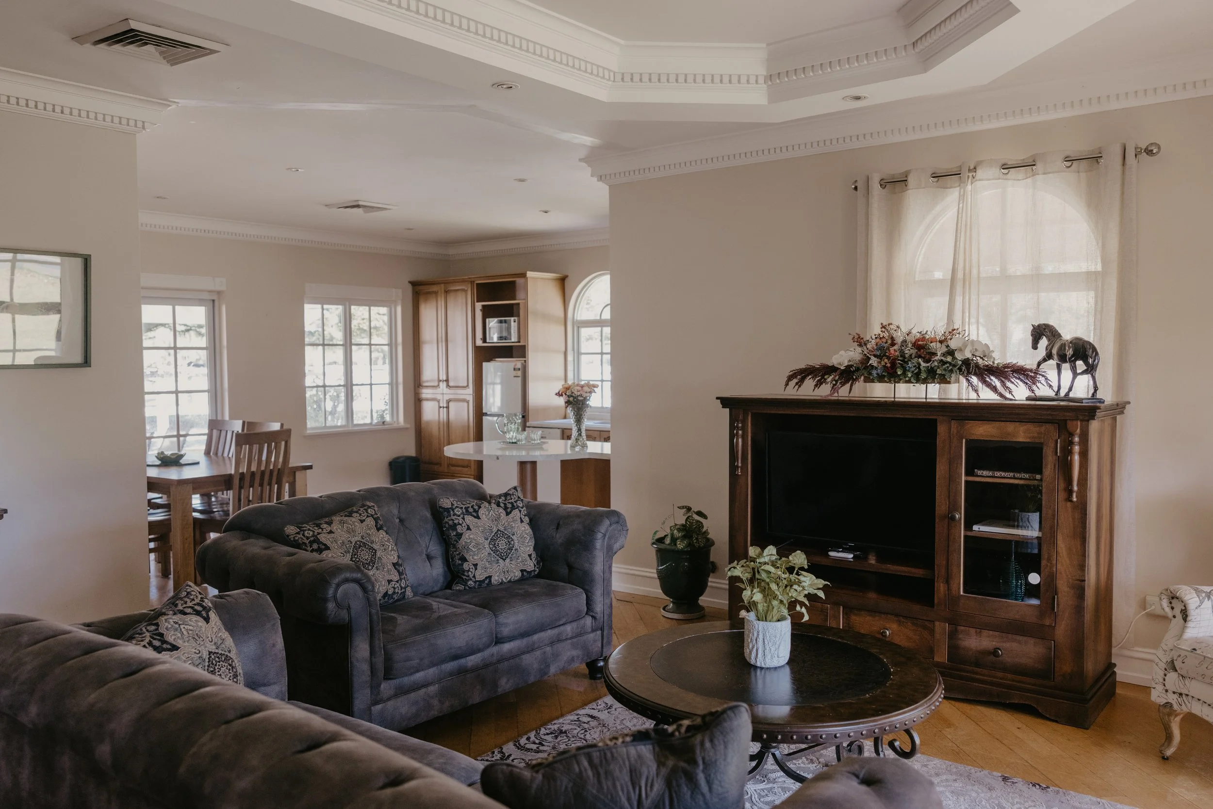Living room with beige walls, large windows with sheer curtains, and wooden furniture, including a television stand with a decorative horse statue and floral arrangement on top, a dining table with chairs, and a gray tufted sofa with patterned cushions.