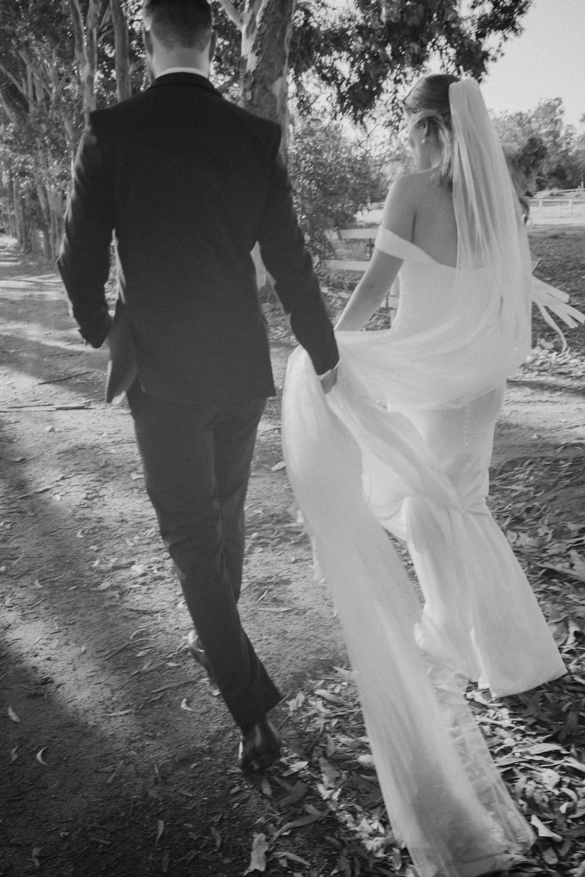 A black and white photo of a bride and groom walking away on a dirt path surrounded by trees and a white fence in the background, with the bride holding her gown.