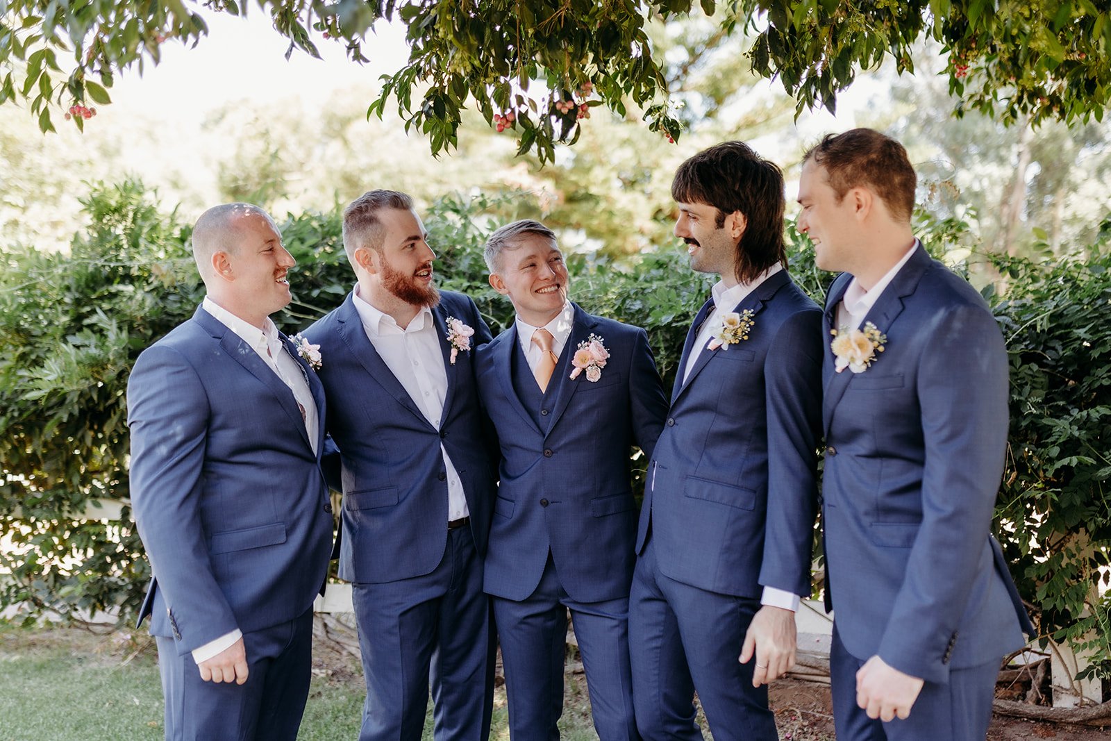 Group of six men in blue suits at a wedding outdoors, smiling and looking at each other, with greenery and trees in the background.