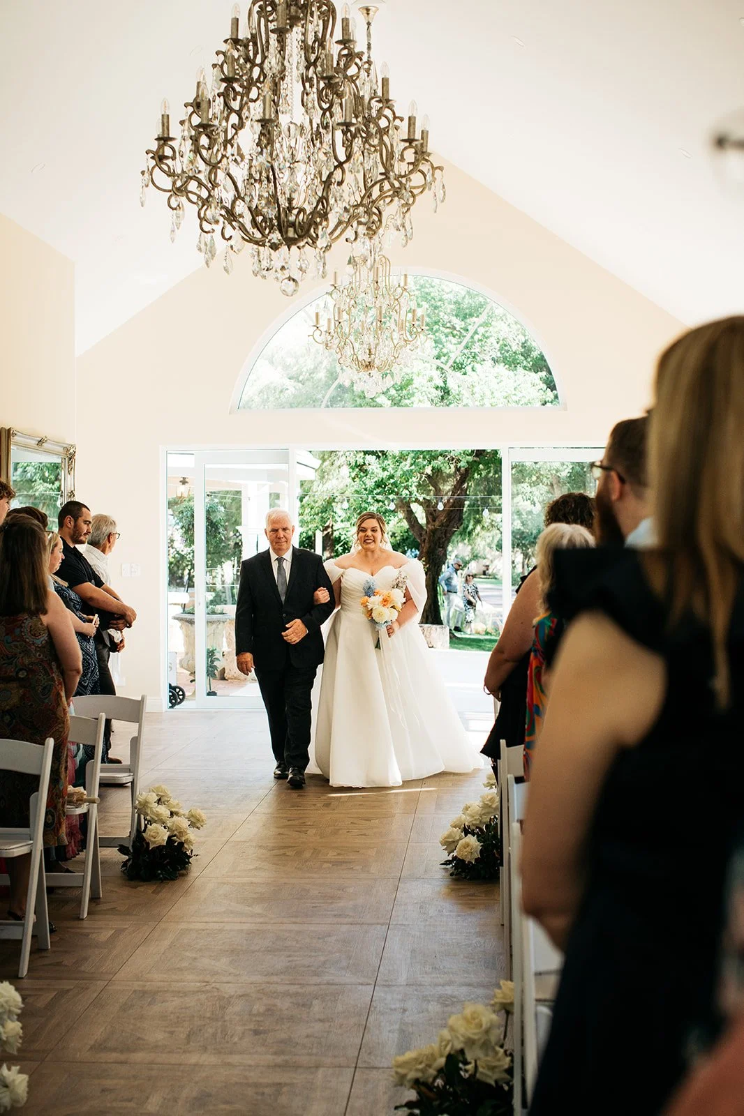 A bride walking down the aisle with her escort, holding a bouquet of flowers inside a bright wedding venue with chandeliers and guests on either side.