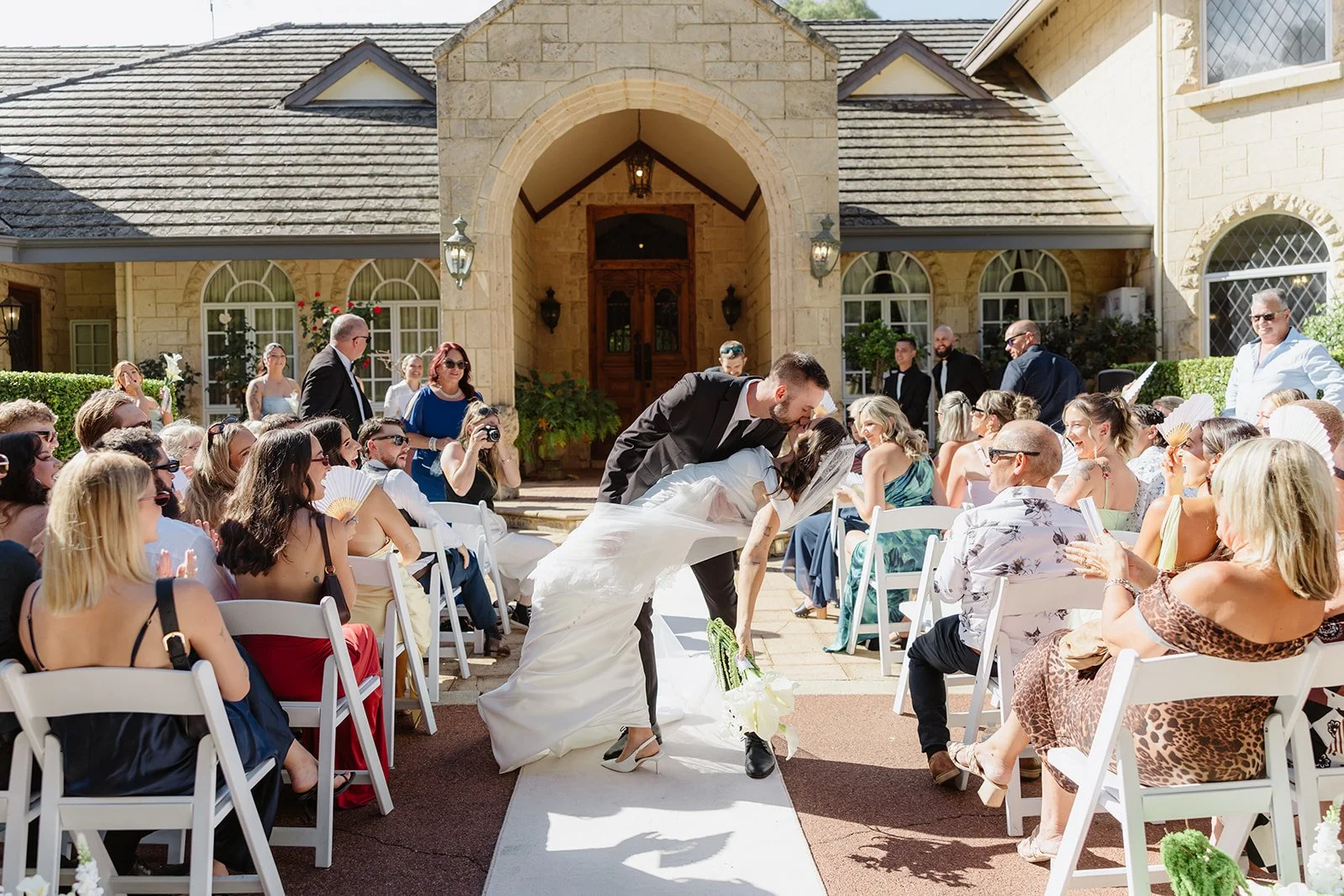 A wedding ceremony taking place outdoors in front of a house with an arched entrance. The bride and groom are kissing, with the bride bending backwards. Guests are seated on white chairs, watching and smiling, some taking photos. The scene is bright 