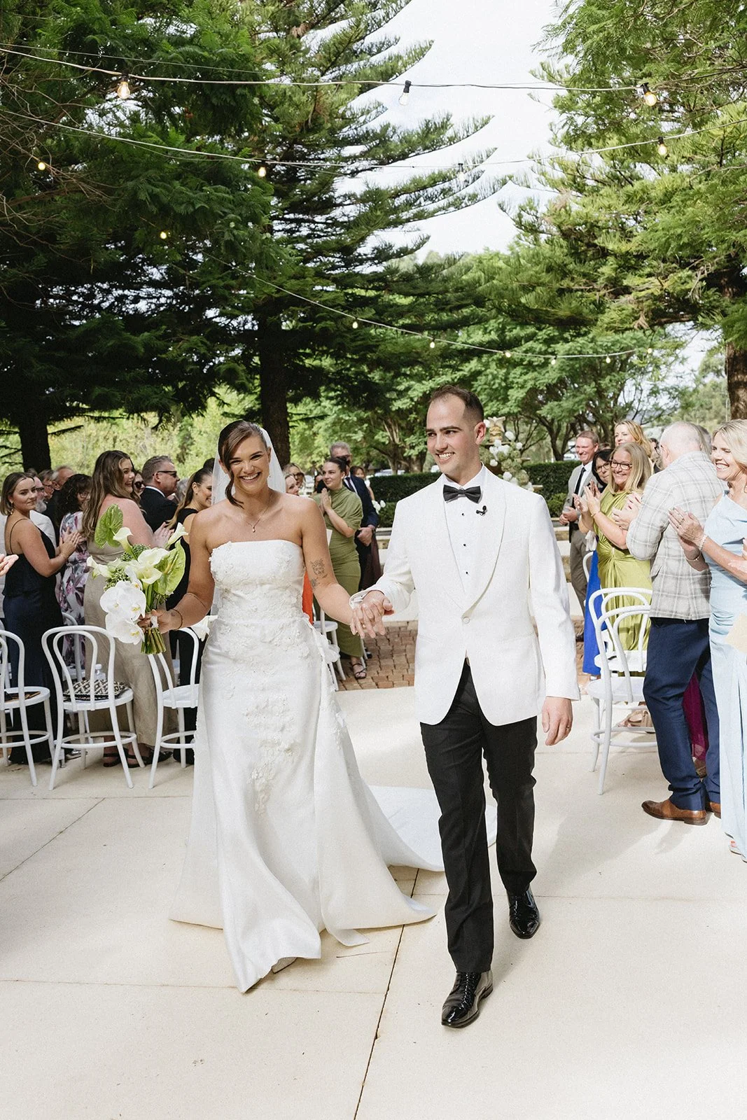 A newlywed couple, the bride in a white strapless wedding gown holding a bouquet, and the groom in a white tuxedo jacket with a bow tie, walking hand in hand down the aisle outdoors. Guests are clapping and celebrating under trees with string lights.