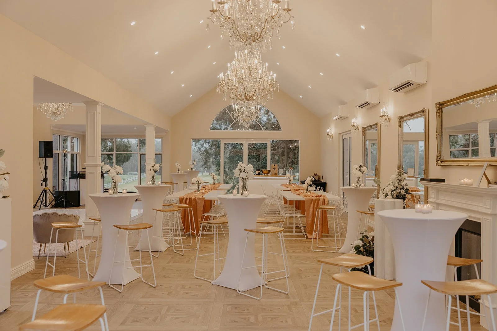 Elegant event space decorated with chandeliers, floral centerpieces, and round cocktail tables with white covers, set up for a celebration.