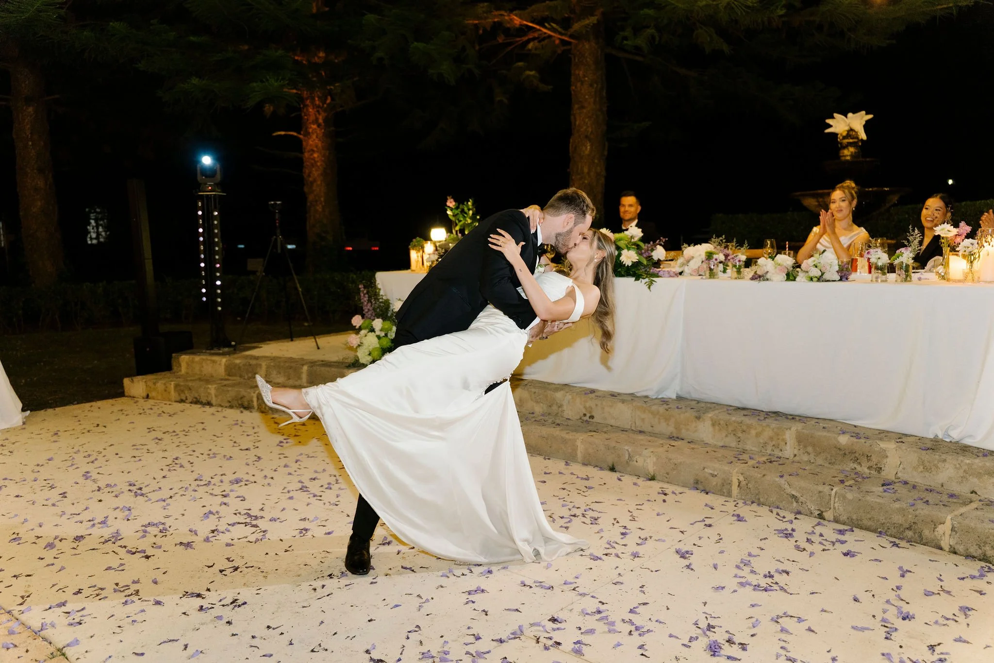 A newlywed couple sharing their first dance at night, with the groom dipping the bride, who is wearing a white wedding gown, while a head table with floral arrangements and two women clapping is visible in the background.
