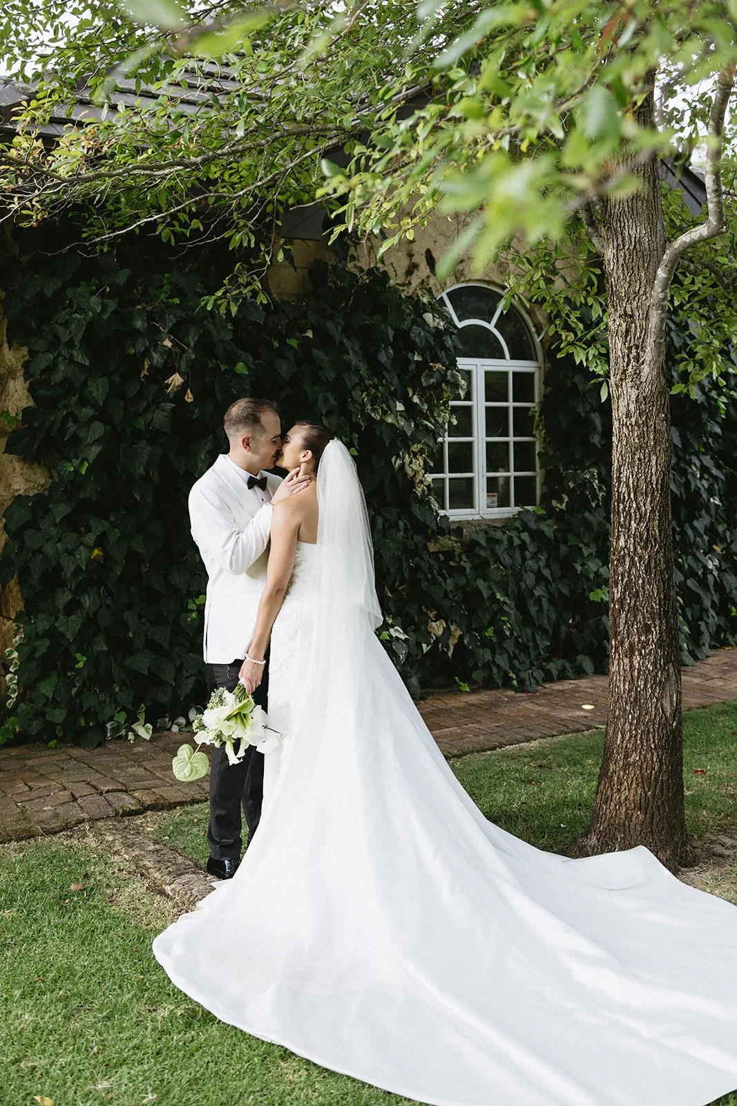 A bride and groom share a kiss outdoors, with the bride holding a bouquet of white flowers and wearing a long white wedding gown and veil. The groom wears a white tuxedo jacket and black pants. The background features green foliage, a brick path, and