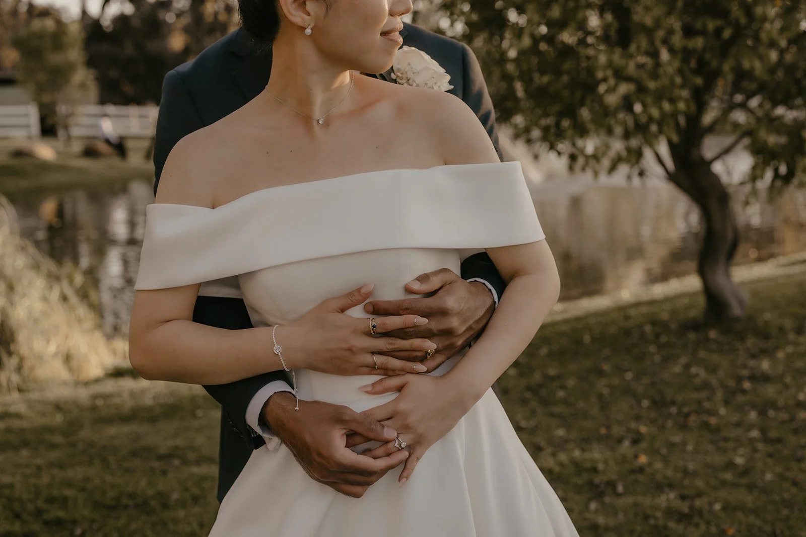 Couple embraces outdoors, woman in off-shoulder white dress, man in dark suit, trees and pond in background.