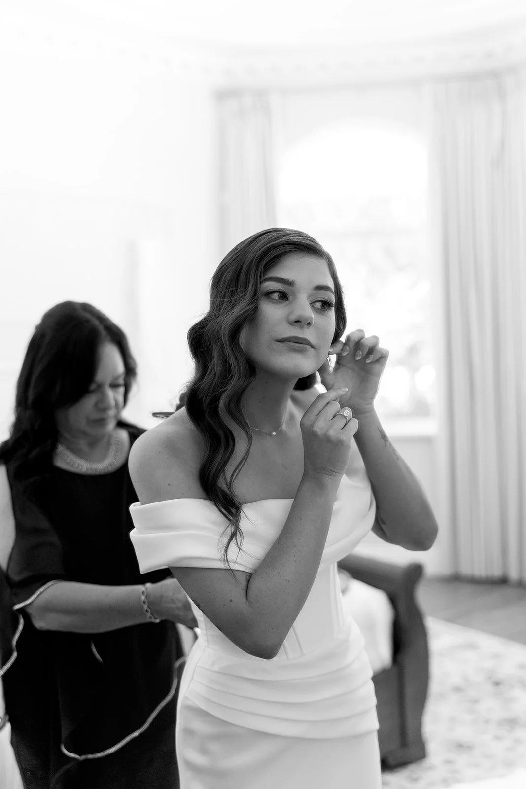 A woman with long wavy hair and an off-shoulder dress adjusts earrings, with another woman in the background.