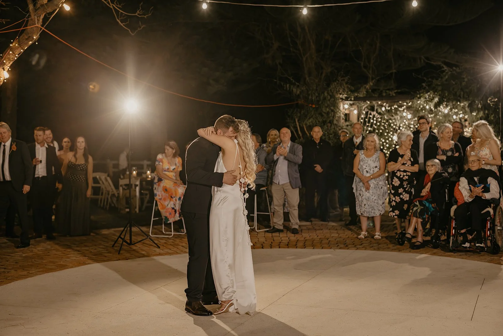 A bride and groom share a dance at their wedding reception outside at night, surrounded by family and friends. String lights and decorative lights are visible in the background in the Swan Valley Perth at Brookleigh Estate