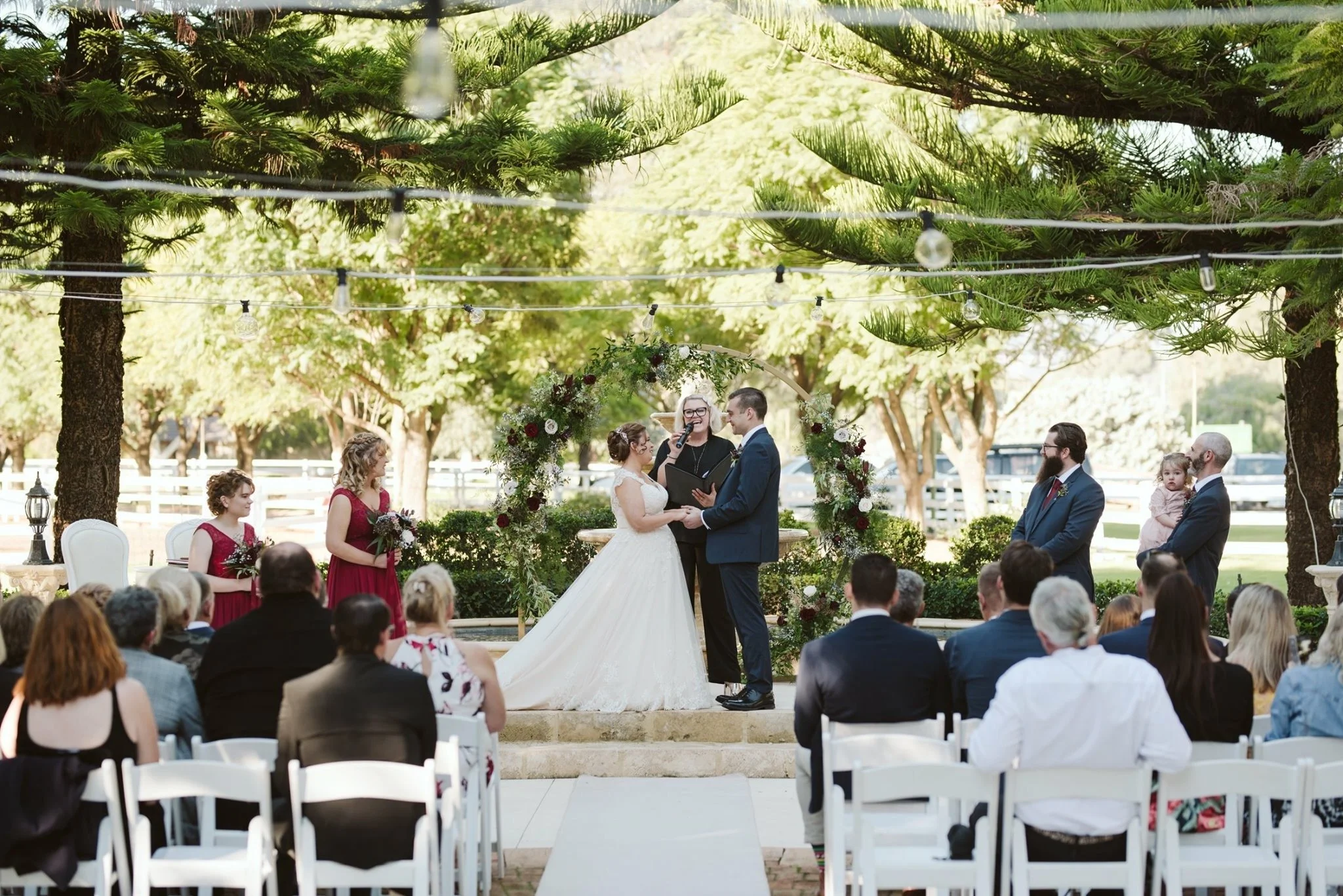 A wedding ceremony taking place outdoors, with a bride and groom holding hands and facing each other beneath a floral arch. Guests are seated in white chairs, and there are trees with string lights hanging above.