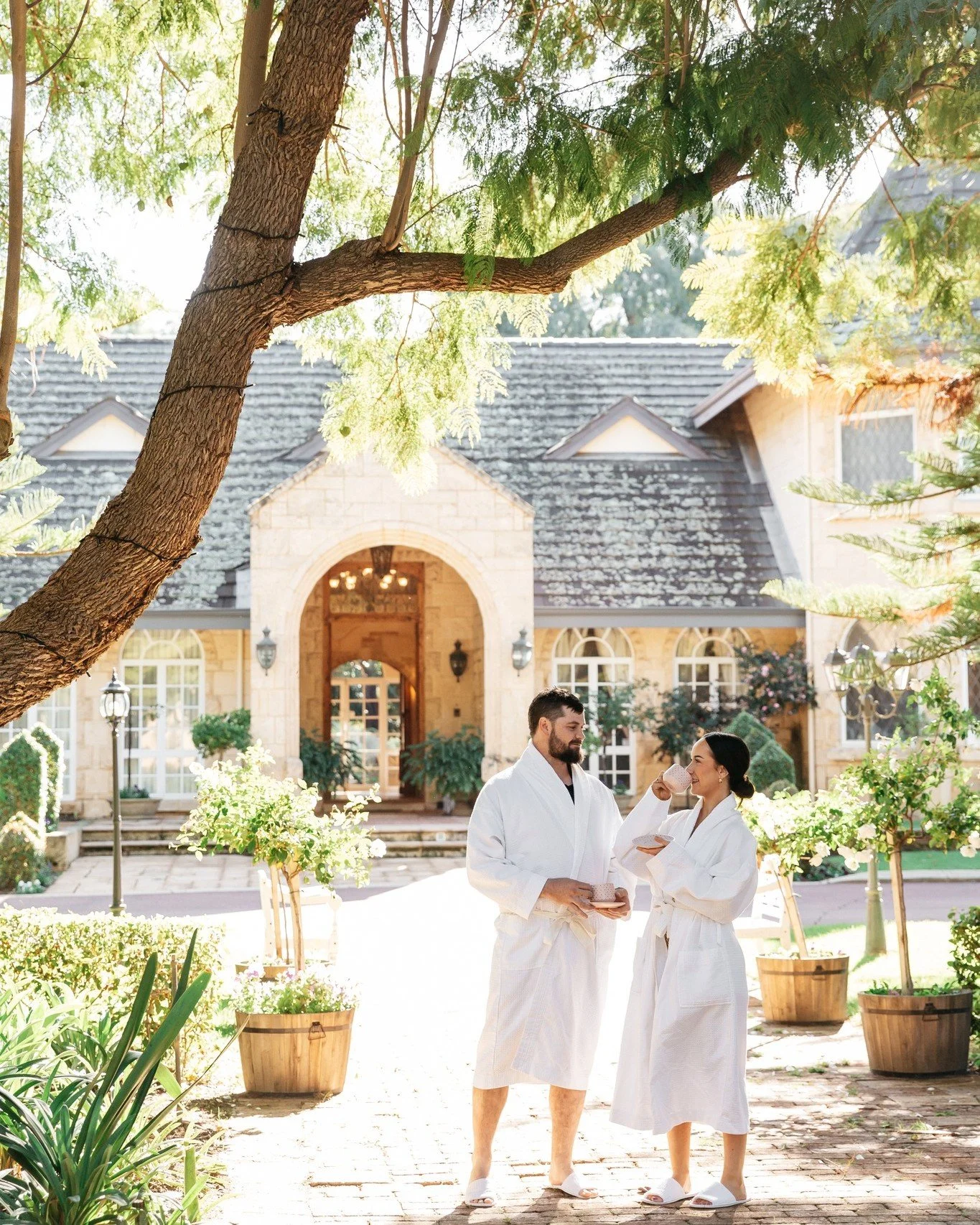 A man and woman in white robes standing outdoors in front of a large stone house, holding cups and a tray, with trees and plants around them.